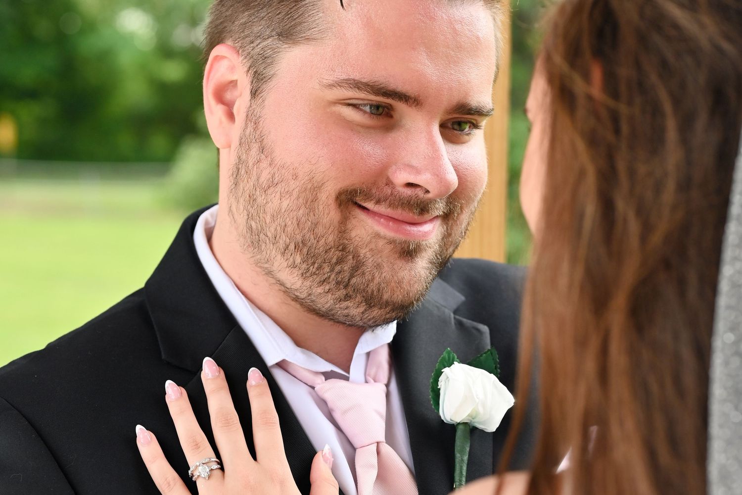 Groom in gray suit with pink tie and white boutonniere smiling at bride during outdoor wedding ceremony.