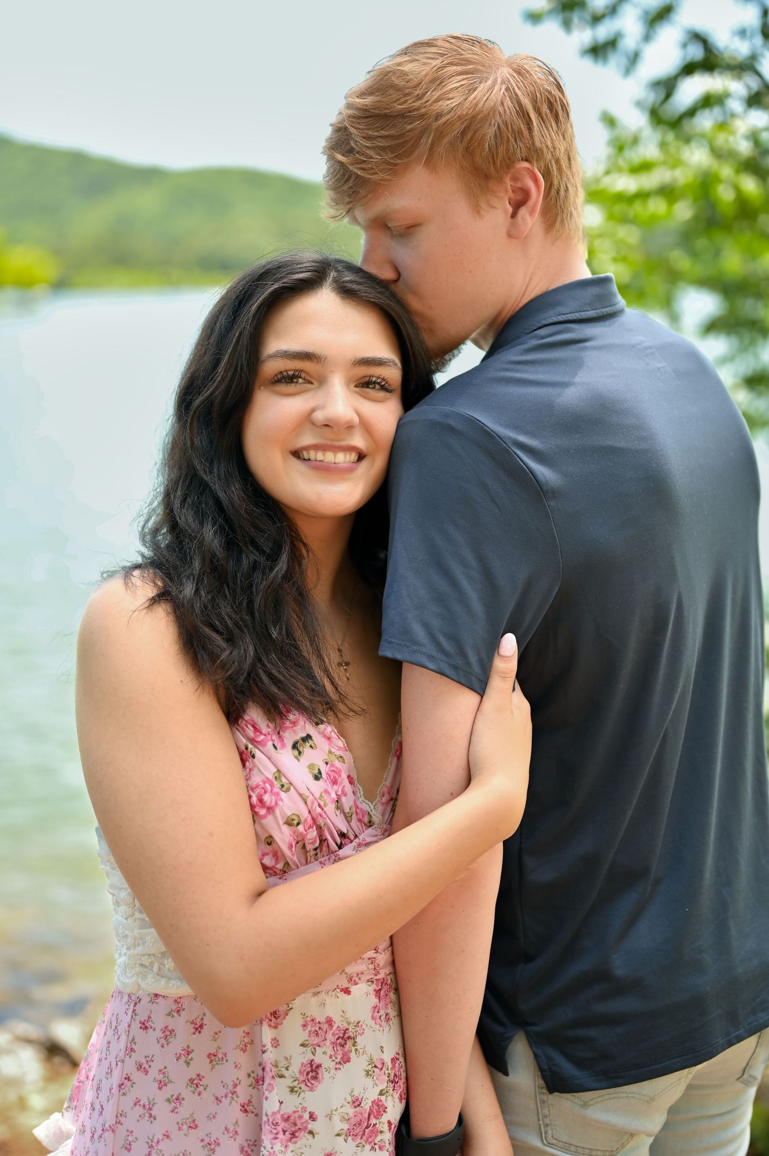 Young couple sharing a tender moment by a lake, man kissing woman's cheek as she smiles at camera.