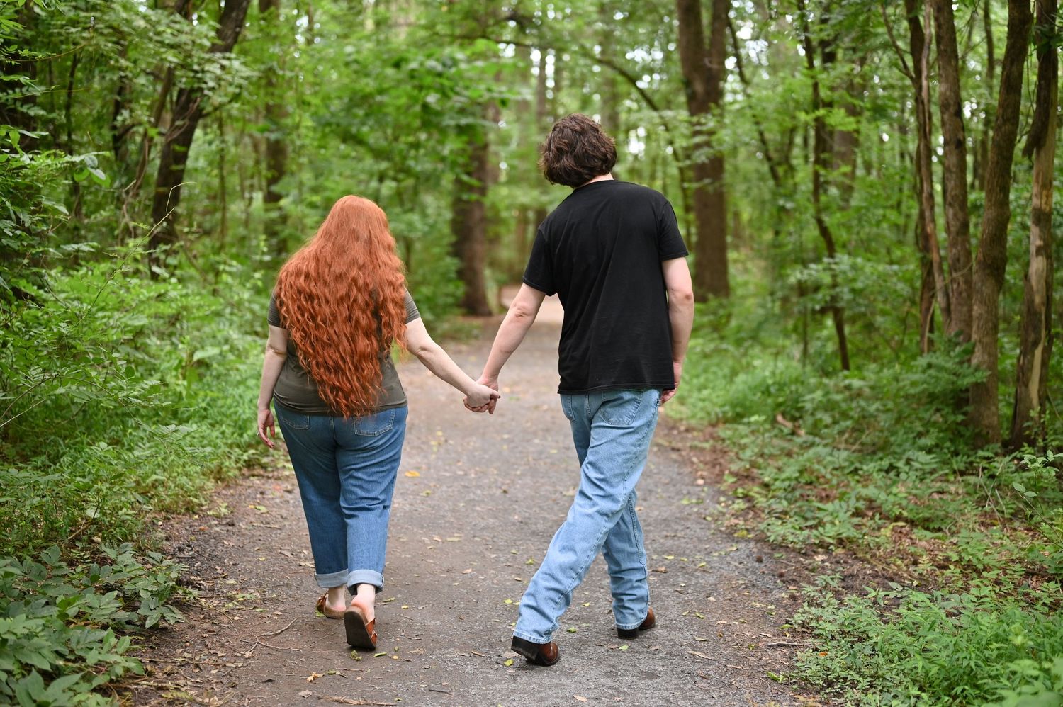 Couple holding hands walking on a forest path surrounded by lush green trees, woman with long red hair.