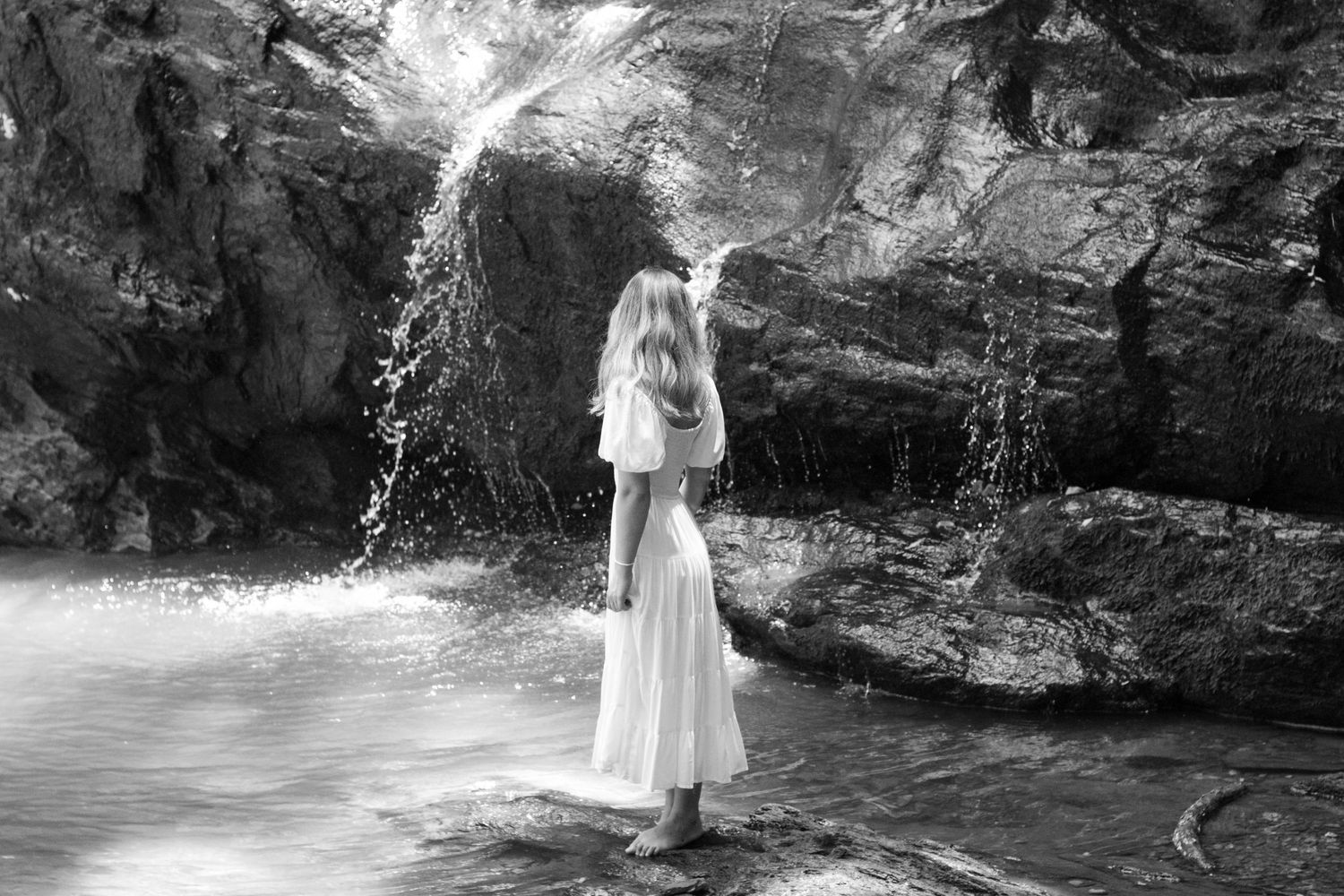 Black and white photo of a woman in a white dress standing near a waterfall in nature.