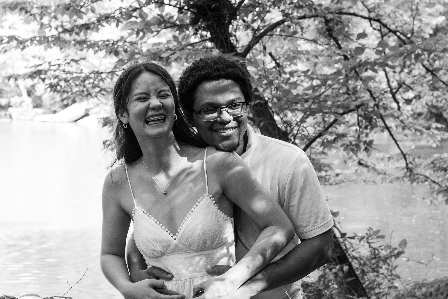 Black and white photo of a smiling couple embracing outdoors near a waterfall and trees.
