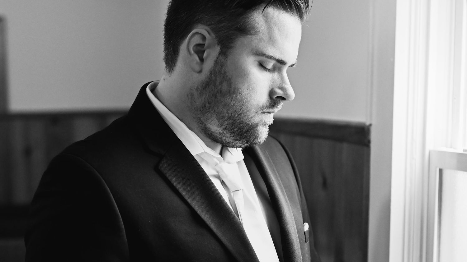 Black and white portrait of a bearded man in a suit looking down near a window.