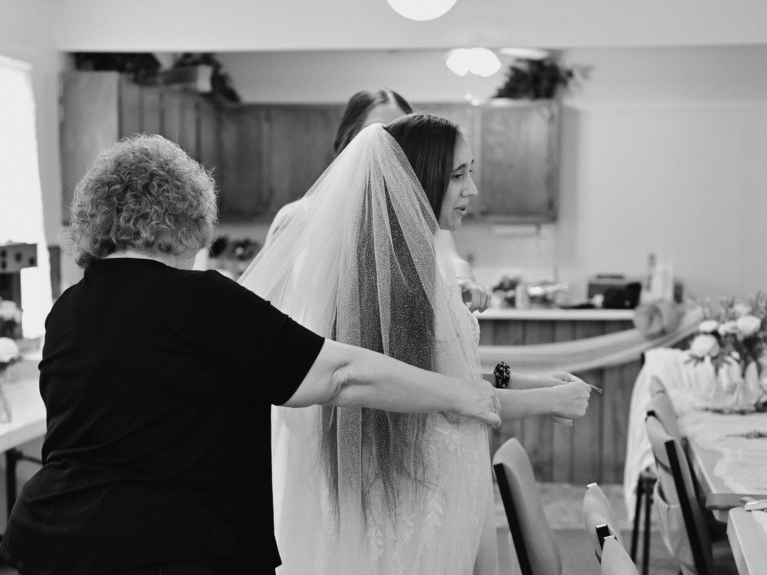 Black and white photo of an older woman helping a bride adjust her veil before the wedding ceremony.