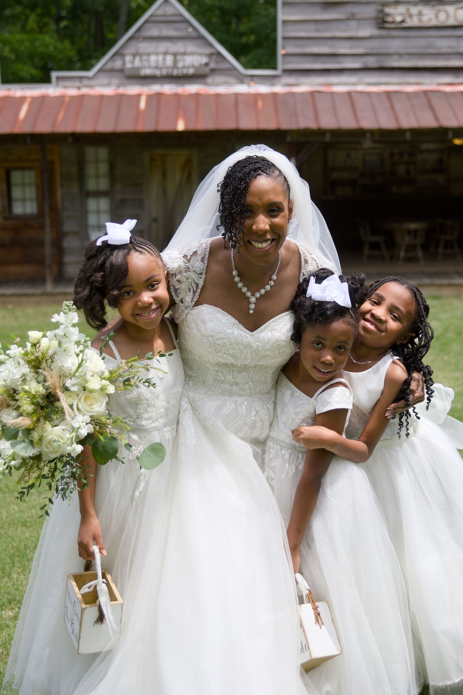 A smiling bride in a white gown and veil poses with three young flower girls in white dresses outdoors.
