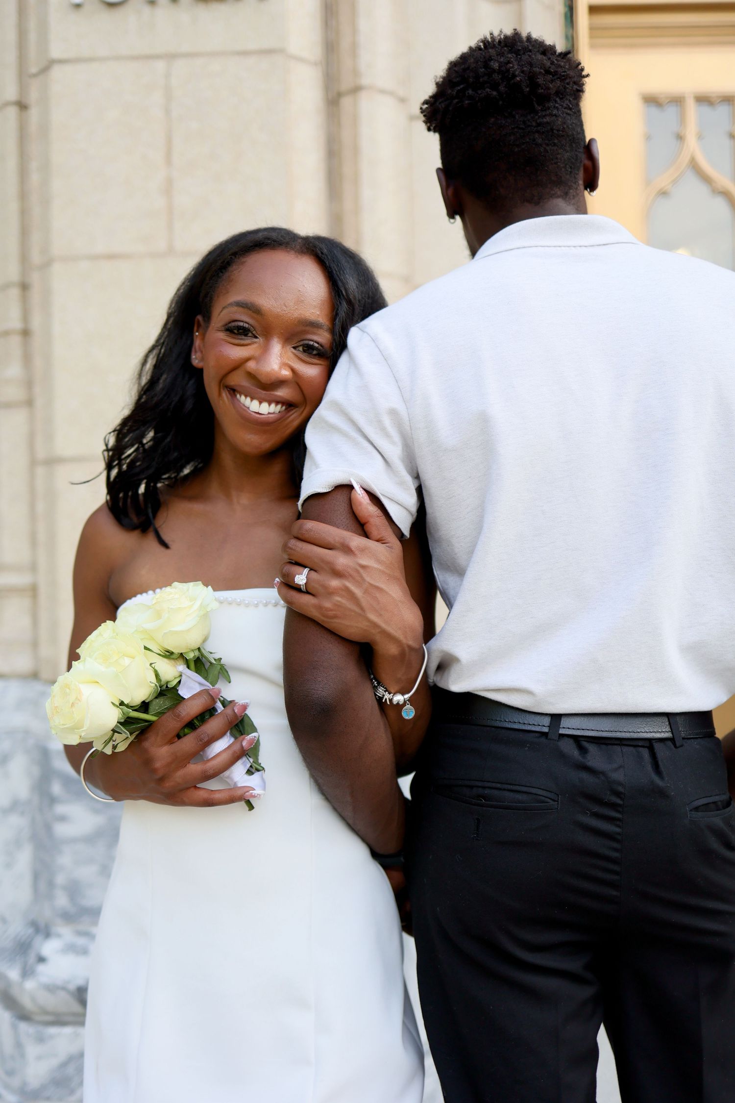 A smiling Black bride in a white strapless gown holds yellow roses while embracing her groom outside a building.