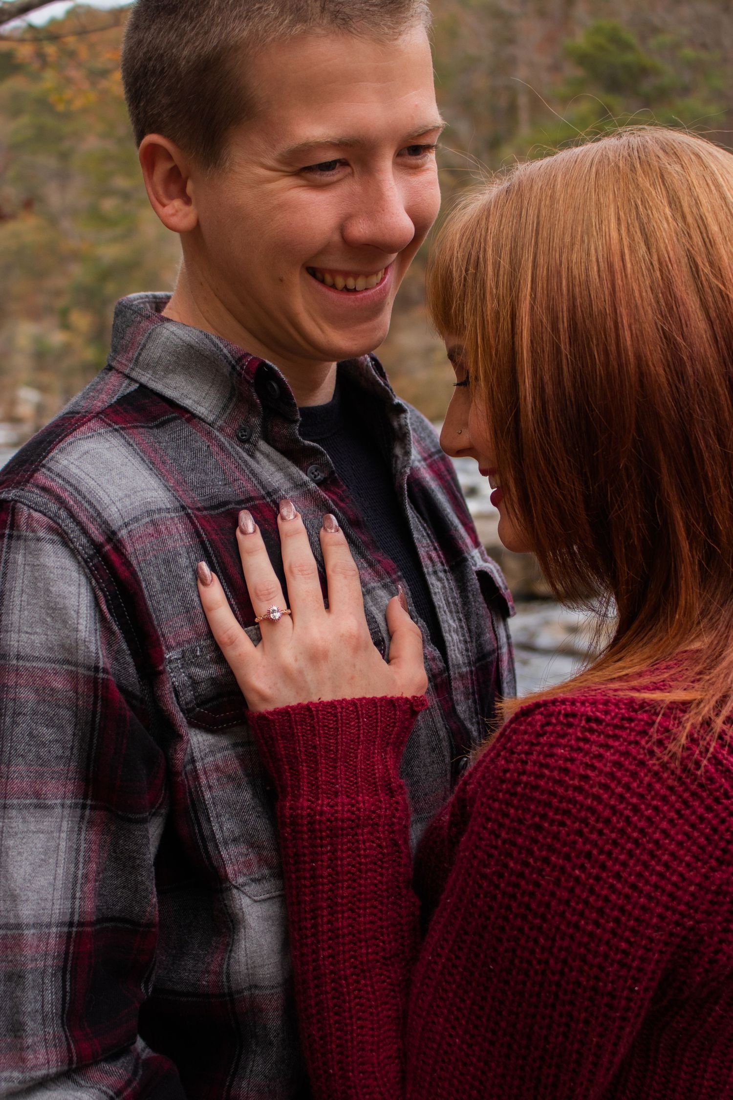 A smiling man in plaid shirt gazes at a redhead woman in burgundy sweater, her hand on his chest showing an engagement ring.