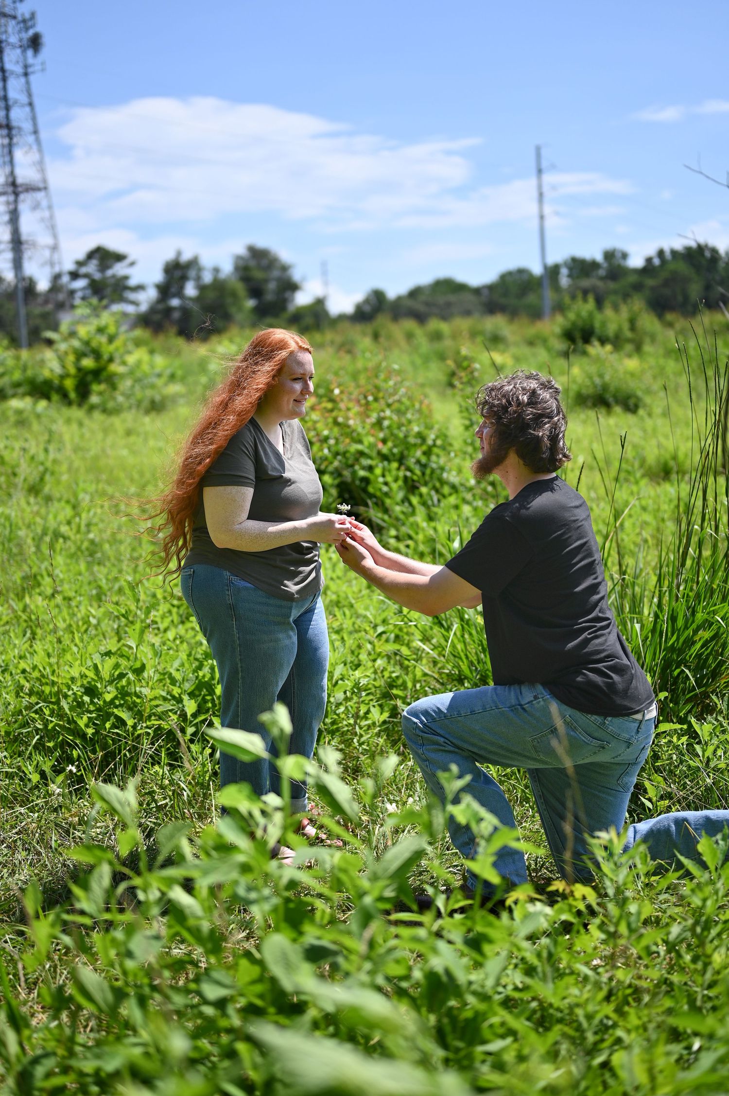 Man on one knee proposing to a red-haired woman in a vibrant green field on a sunny day.