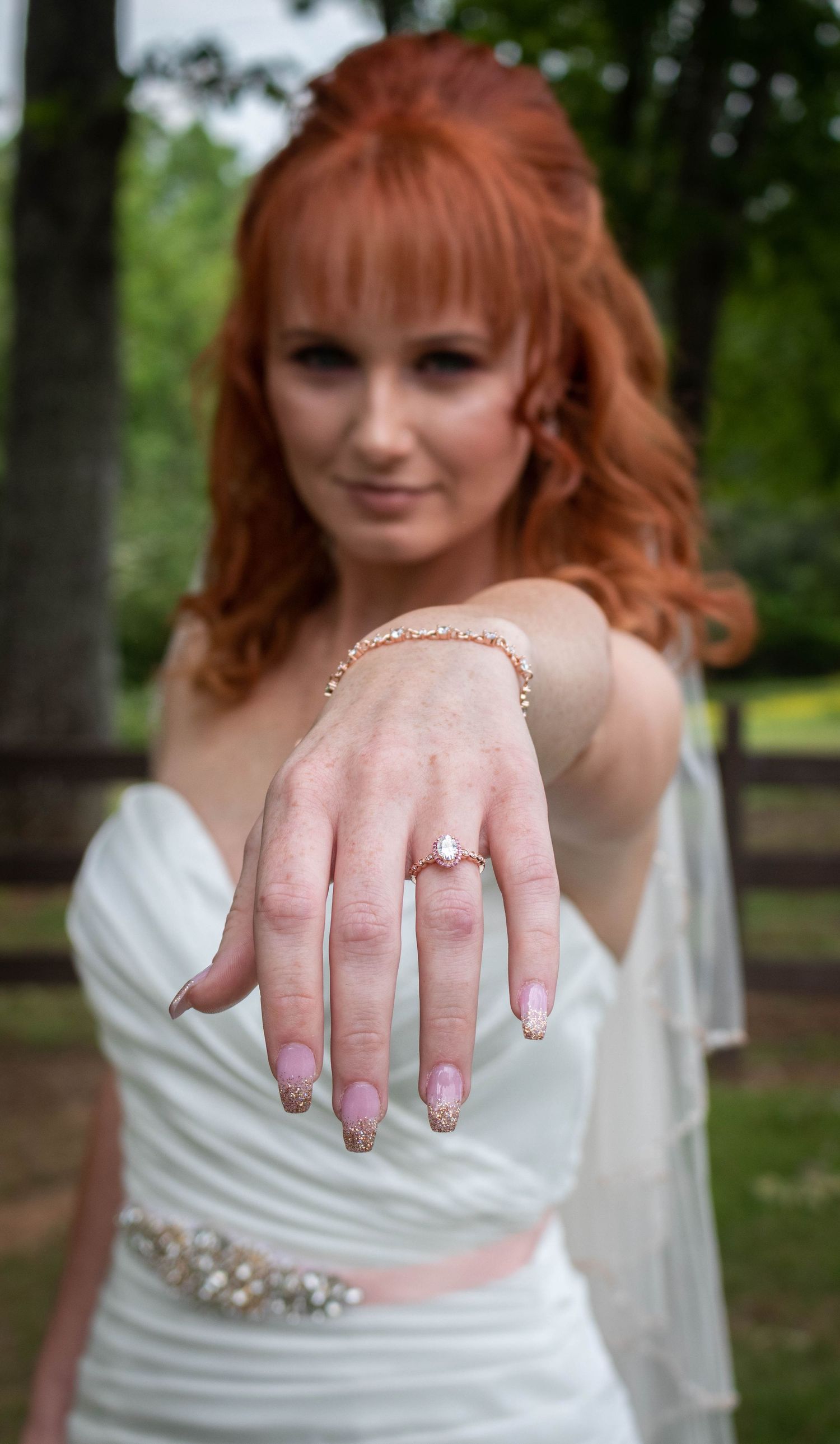 Redhead bride in white gown showing oval diamond engagement ring toward camera outdoors.