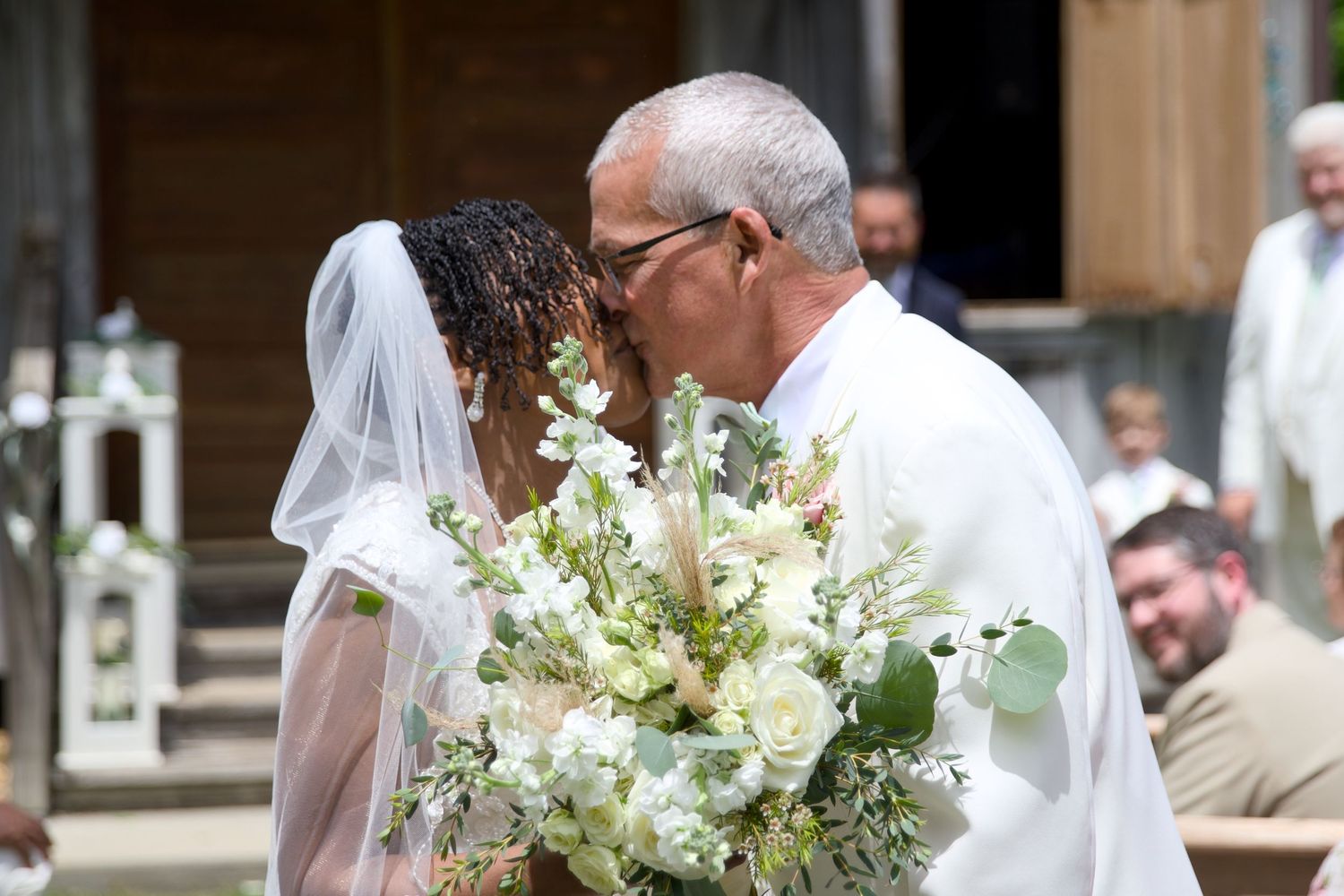 Bride and groom sharing a kiss at outdoor wedding ceremony, bride holding white floral bouquet with greenery.