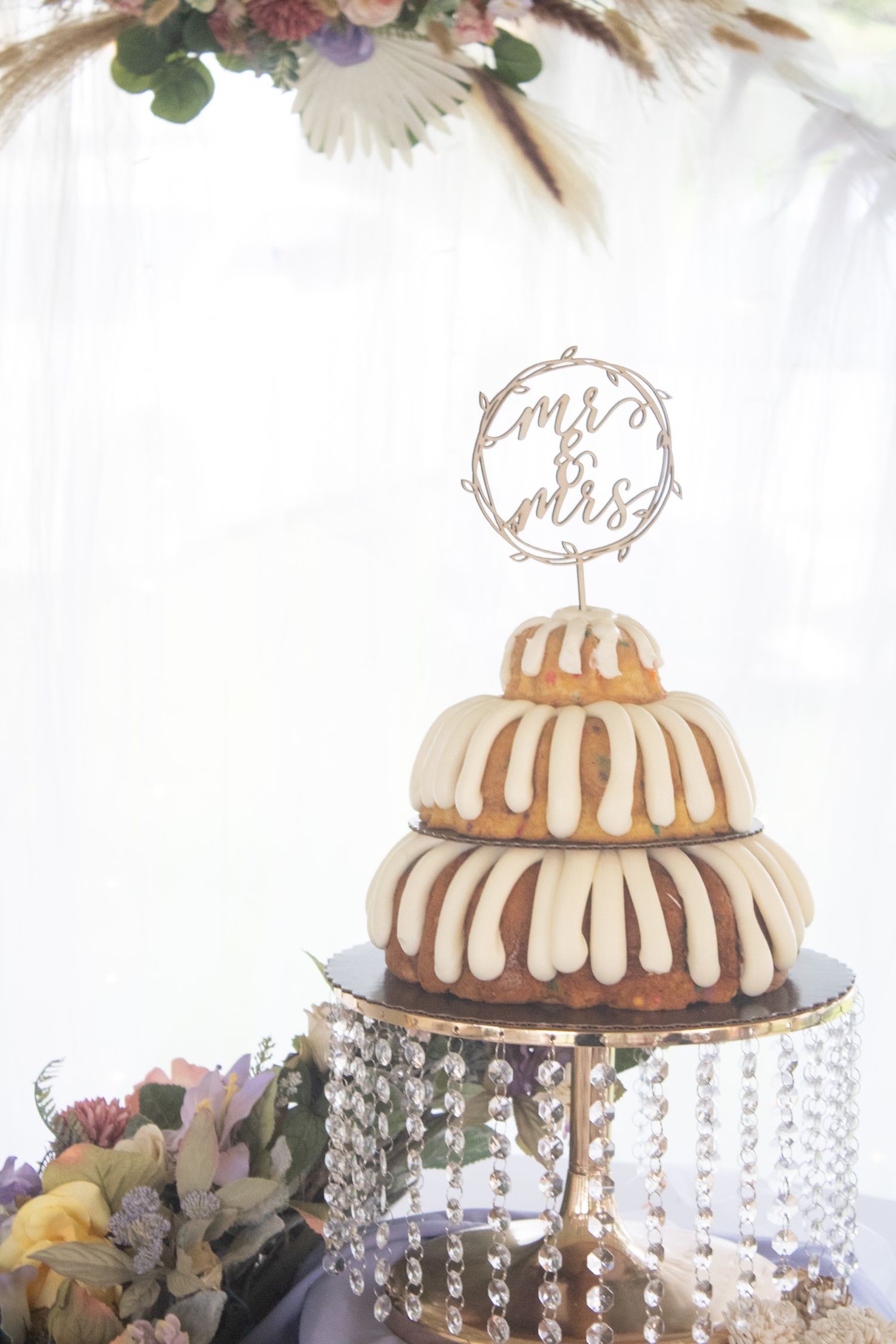 Two-tier bundt wedding cake with white glaze on crystal stand, topped with Mr & Mrs cake topper.