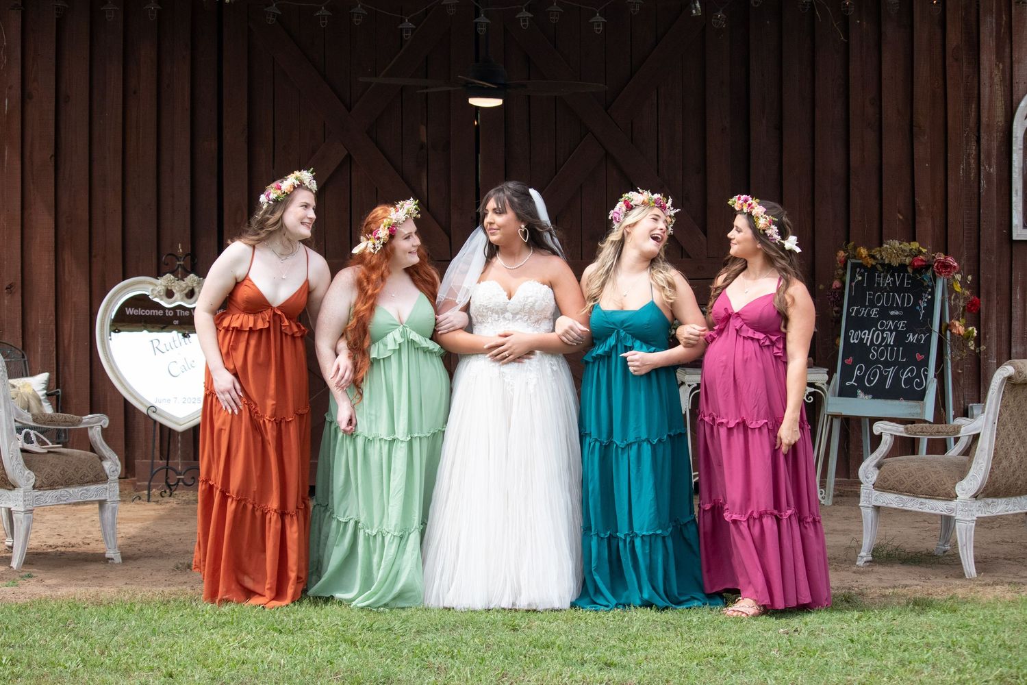 Bride in white gown poses with four bridesmaids in colorful dresses outside rustic barn.