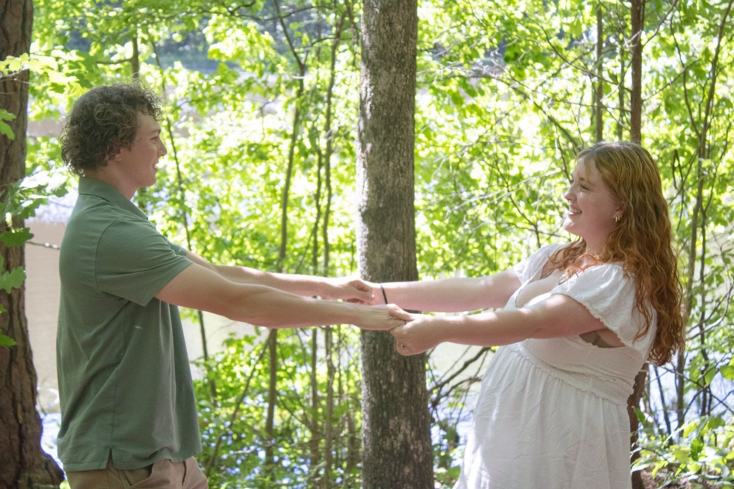 A couple holds hands in a sunlit forest, the woman in white and man in green sharing a romantic outdoor moment.