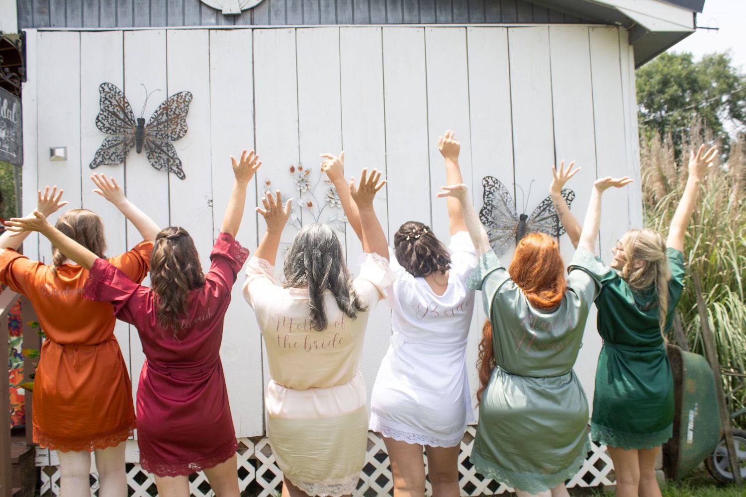 Bride and bridesmaids in colorful robes raising arms together in celebration against white barn wall.
