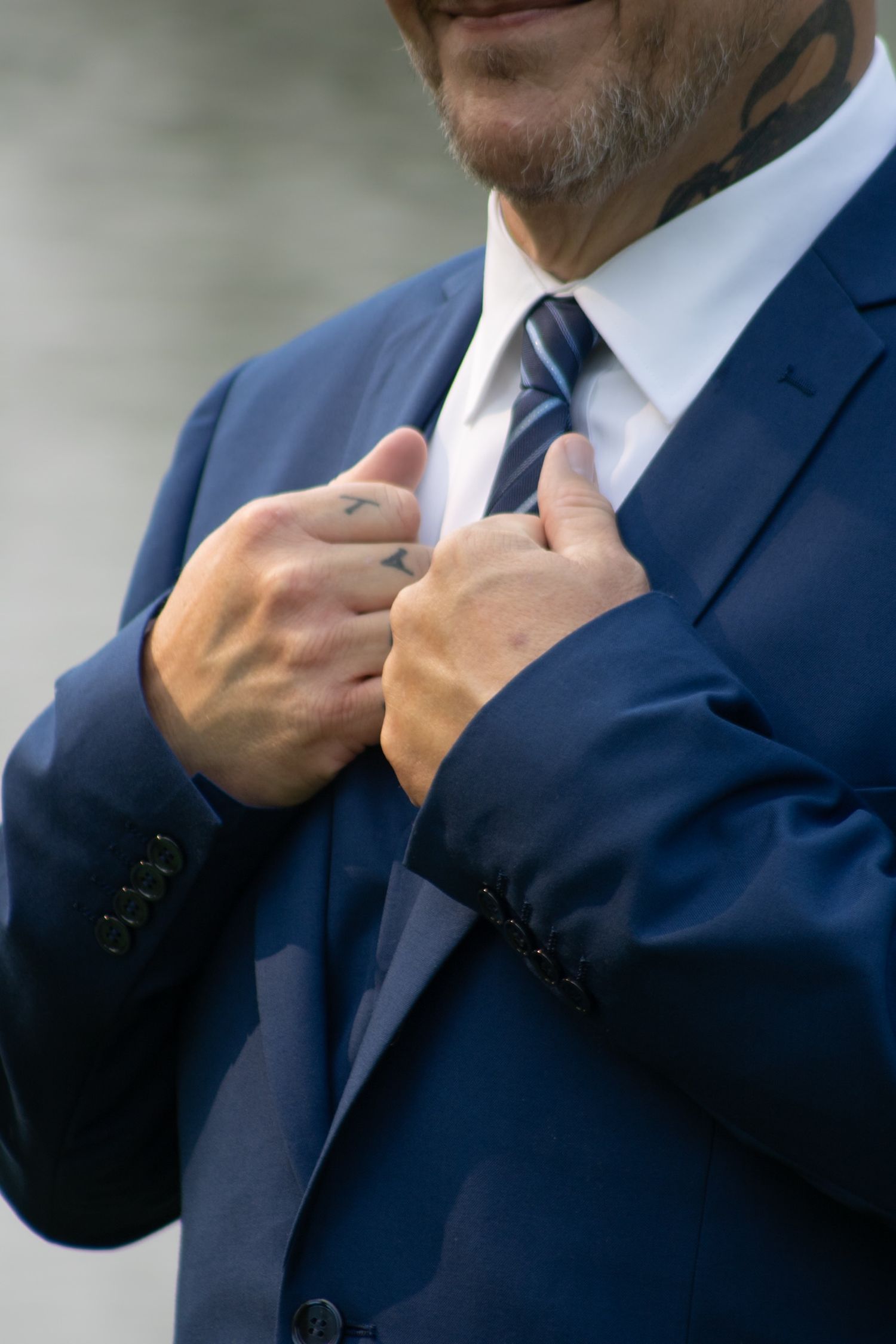 Tattooed man in blue suit adjusting navy striped tie, showing hand and neck tattoos.