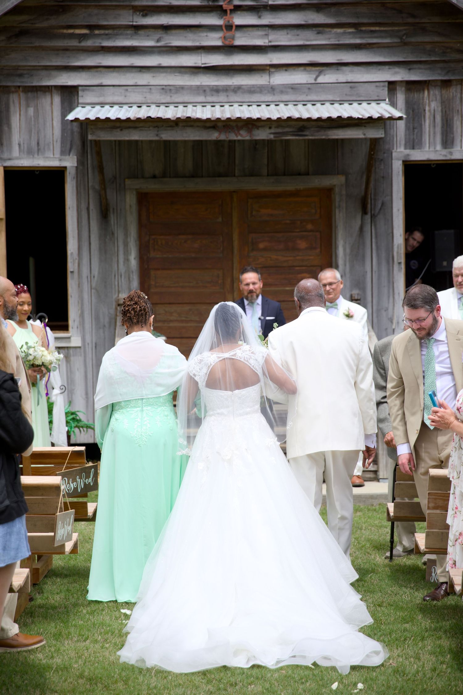 Bride in white gown and veil walks down outdoor aisle toward groom at rustic barn wedding ceremony.