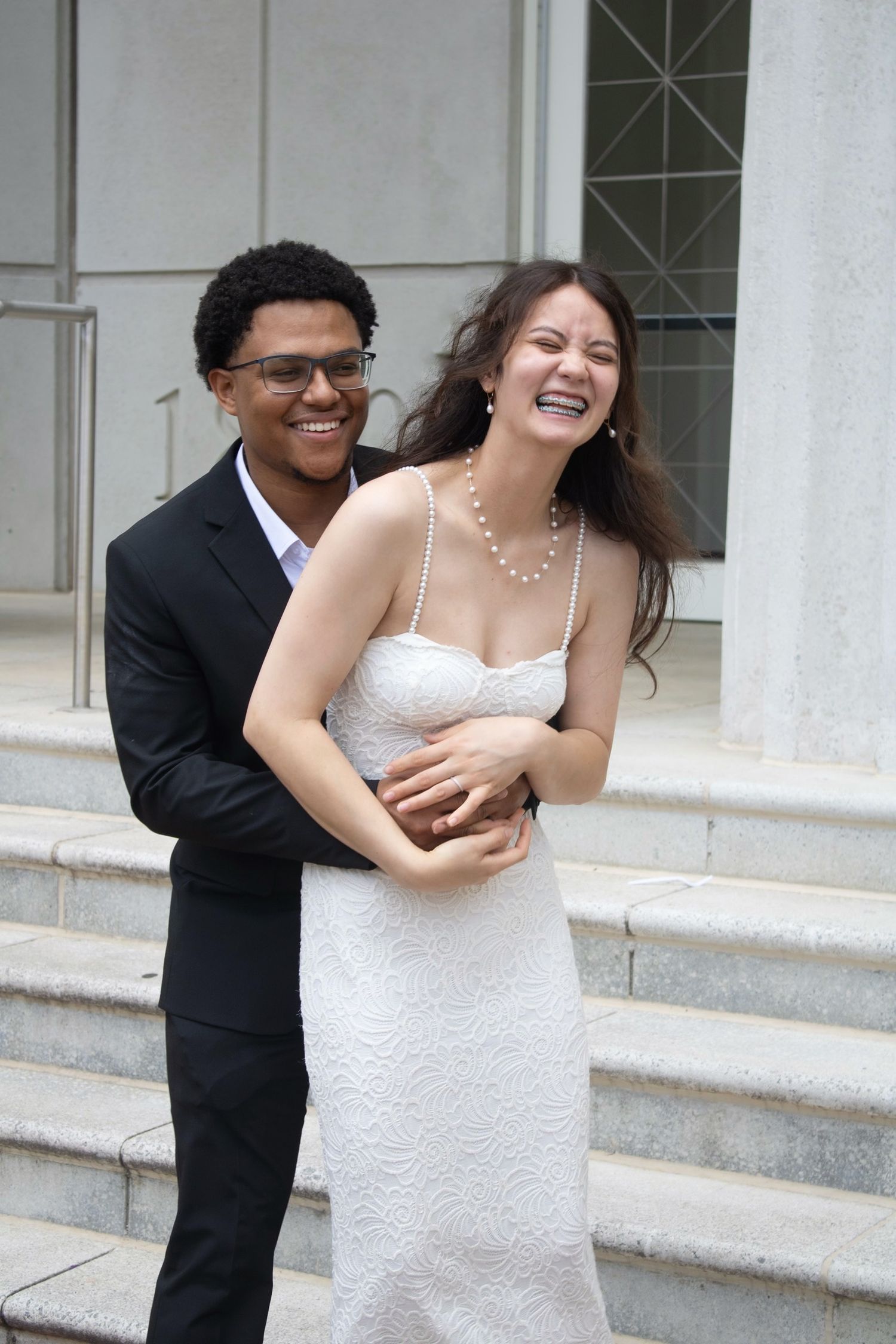 A joyful interracial couple in wedding attire laughing on stone steps, bride in white lace dress with pearl necklace.