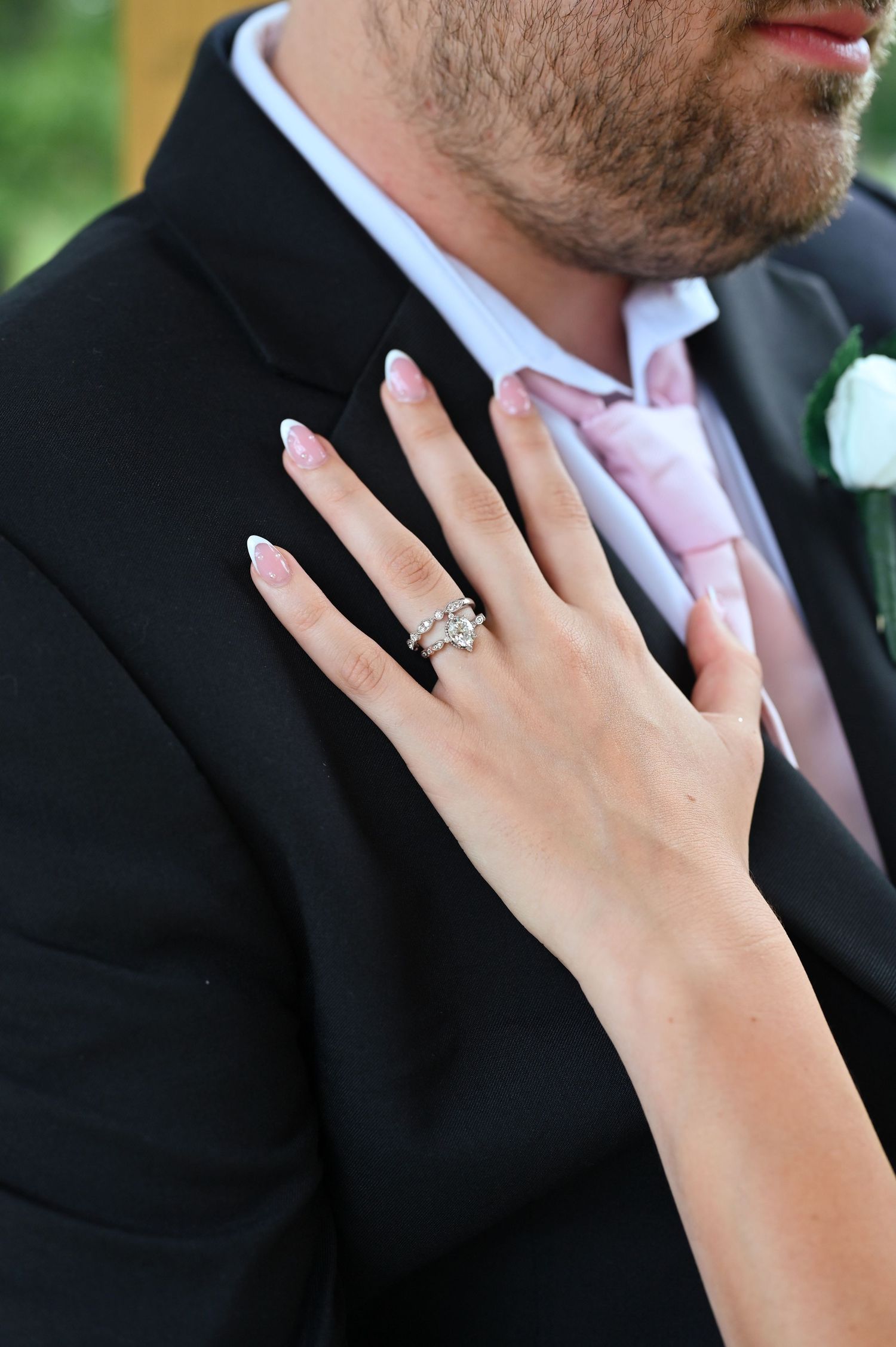 Close-up of bride's hand with diamond wedding ring resting on groom's black tuxedo jacket with pink tie.