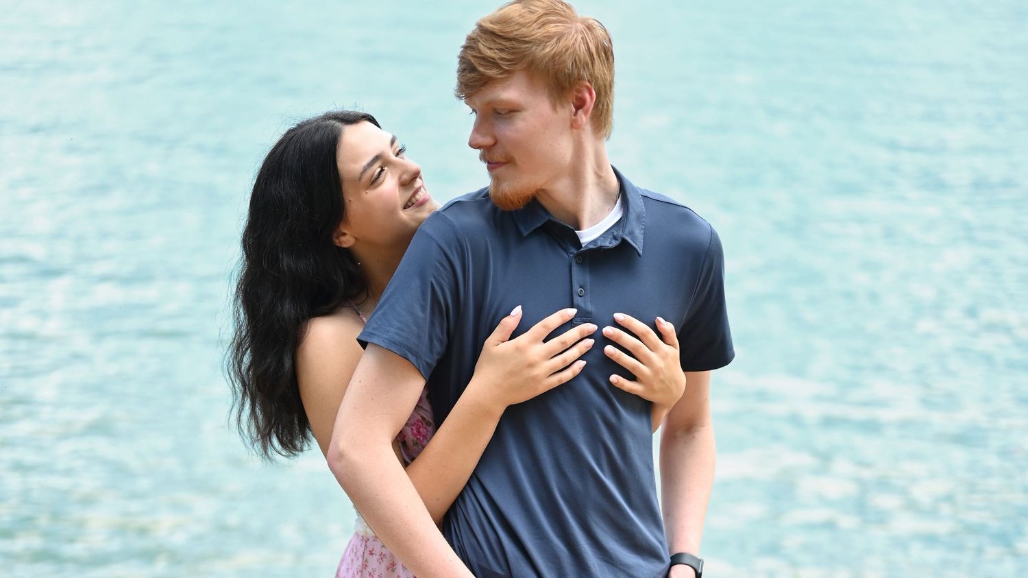 Happy couple embracing closely in front of a bright turquoise lake on a sunny day.