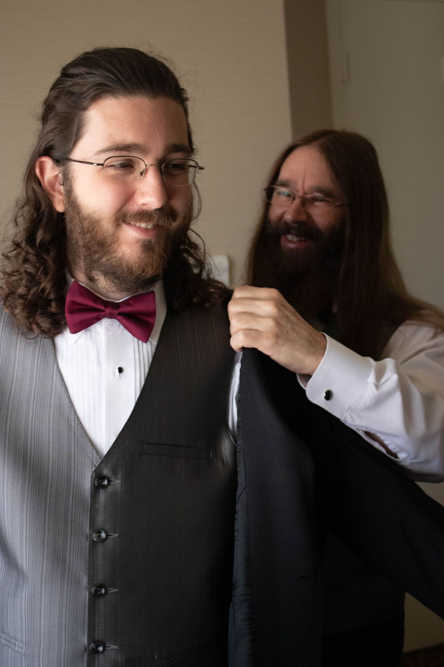 Smiling groom with long hair and burgundy bow tie being helped into jacket by bearded man.