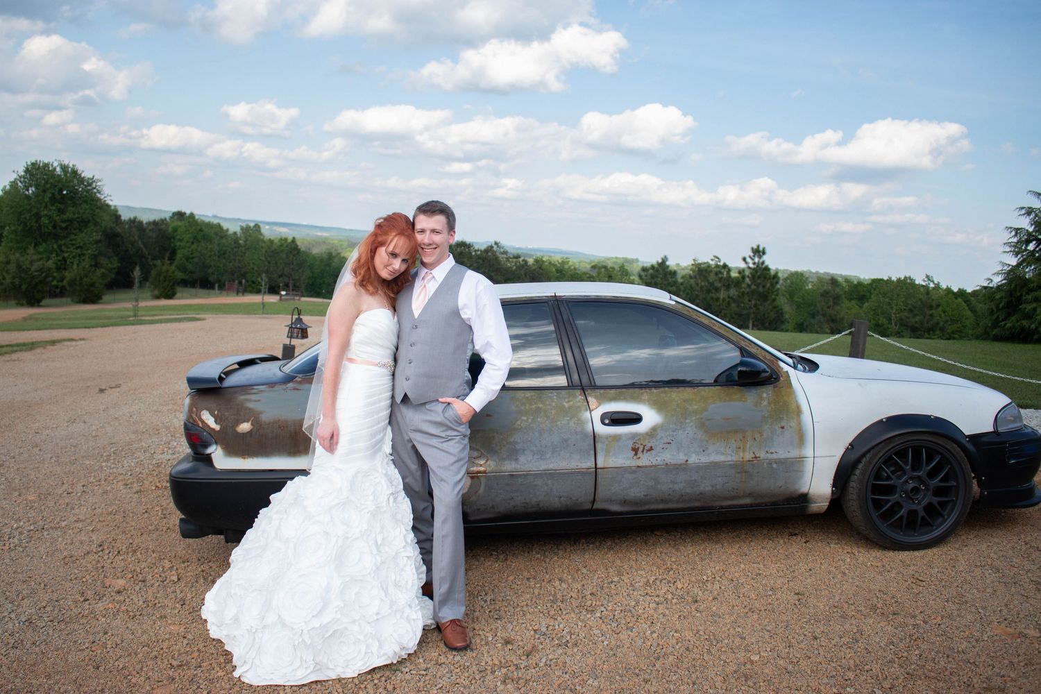 Bride and groom posing together beside vintage rustic car on gravel road.