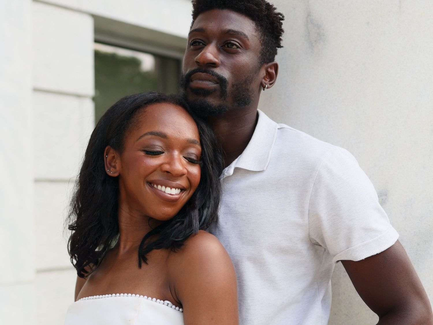 A smiling couple in white outfits pose together outdoors, the woman leaning back against the man.
