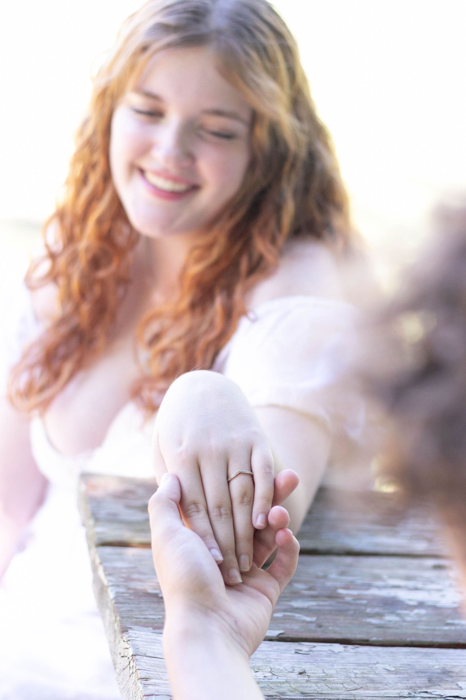 Smiling redhead bride showing off her engagement ring while leaning on a wooden dock.