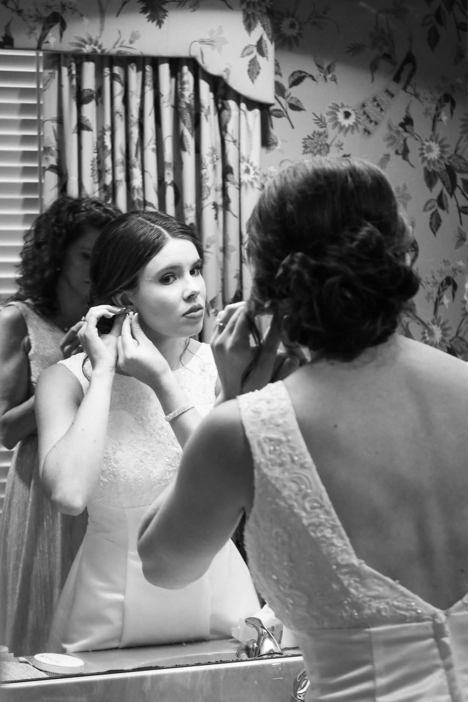 Black and white photo of bride putting on earrings while looking in mirror before wedding ceremony.