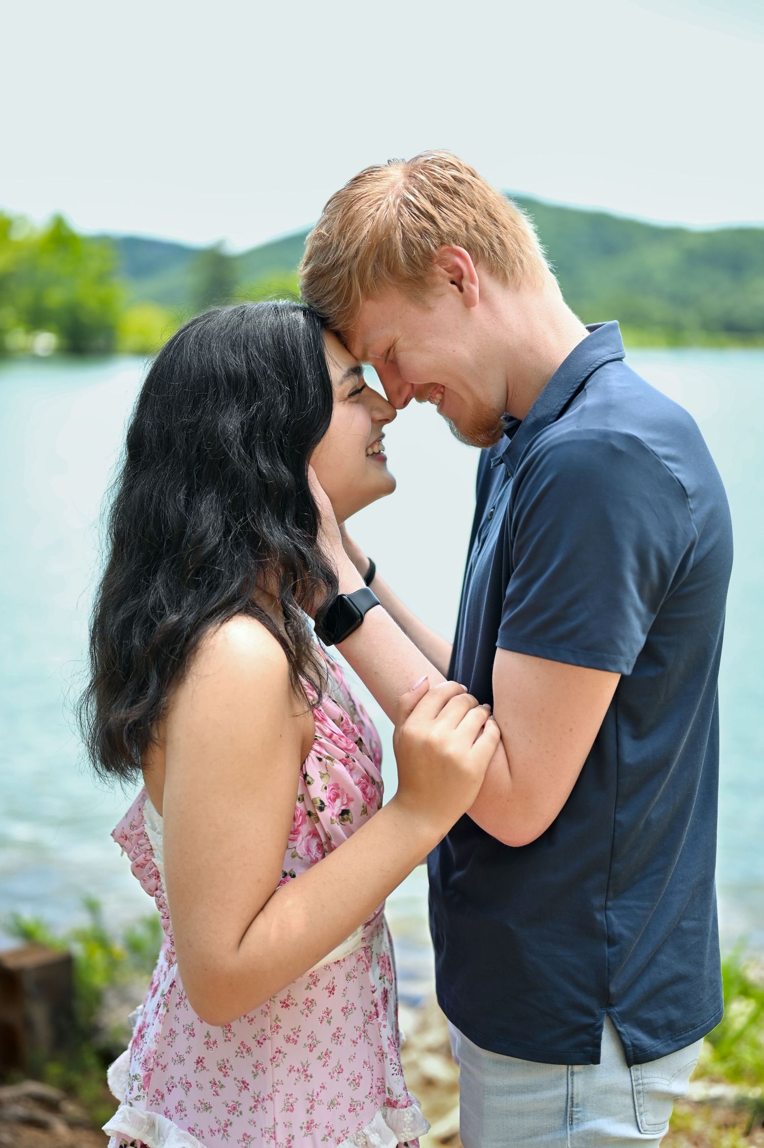 A smiling young couple touches foreheads romantically near a lake, the woman in a floral dress holding the mans arm.