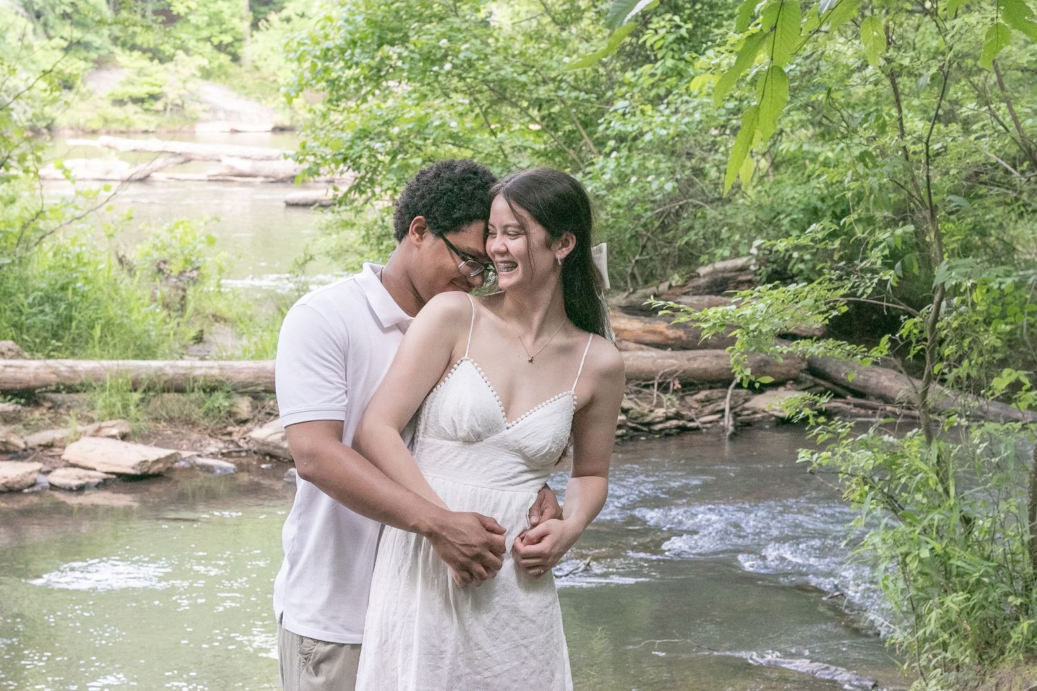Couple embracing by a serene forest creek, woman in white dress smiling as man holds her from behind.