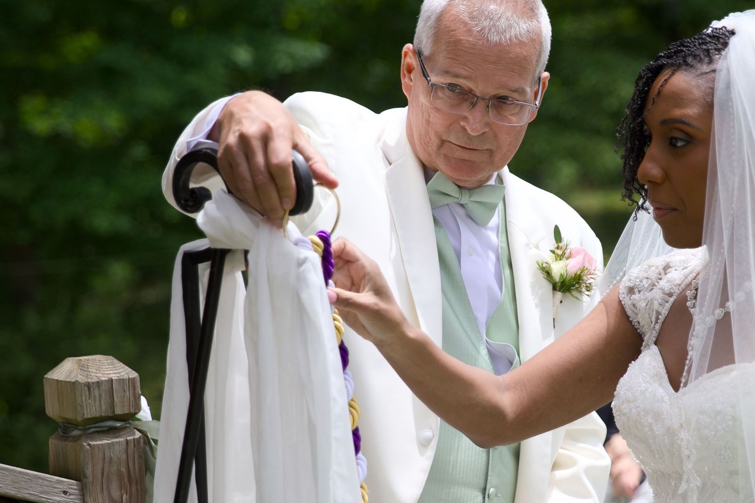 Officiant in white robe assists bride in wedding dress during outdoor ceremony ritual.