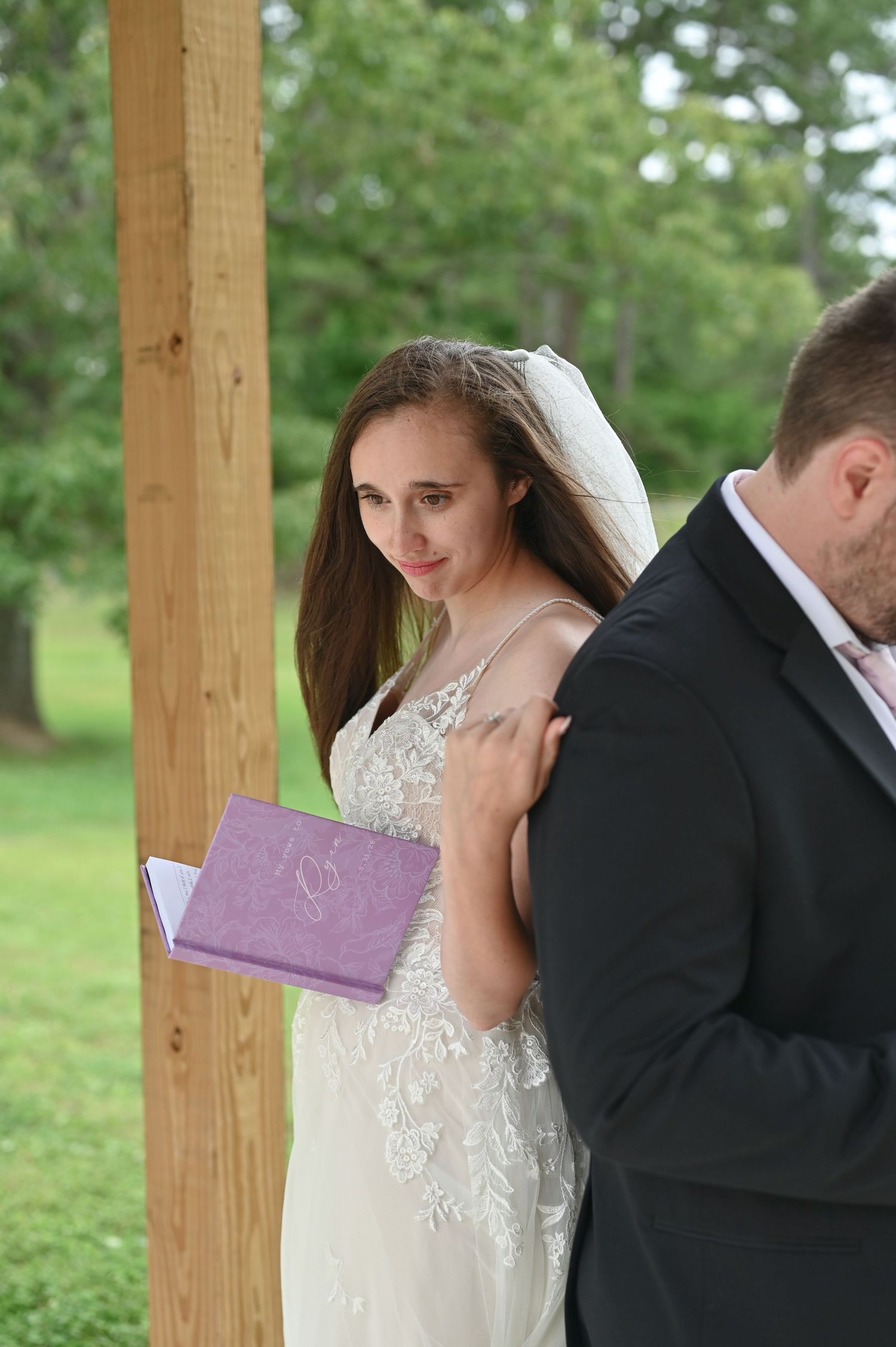 Bride in white lace gown holding purple book stands beside groom in black suit during outdoor wedding ceremony.