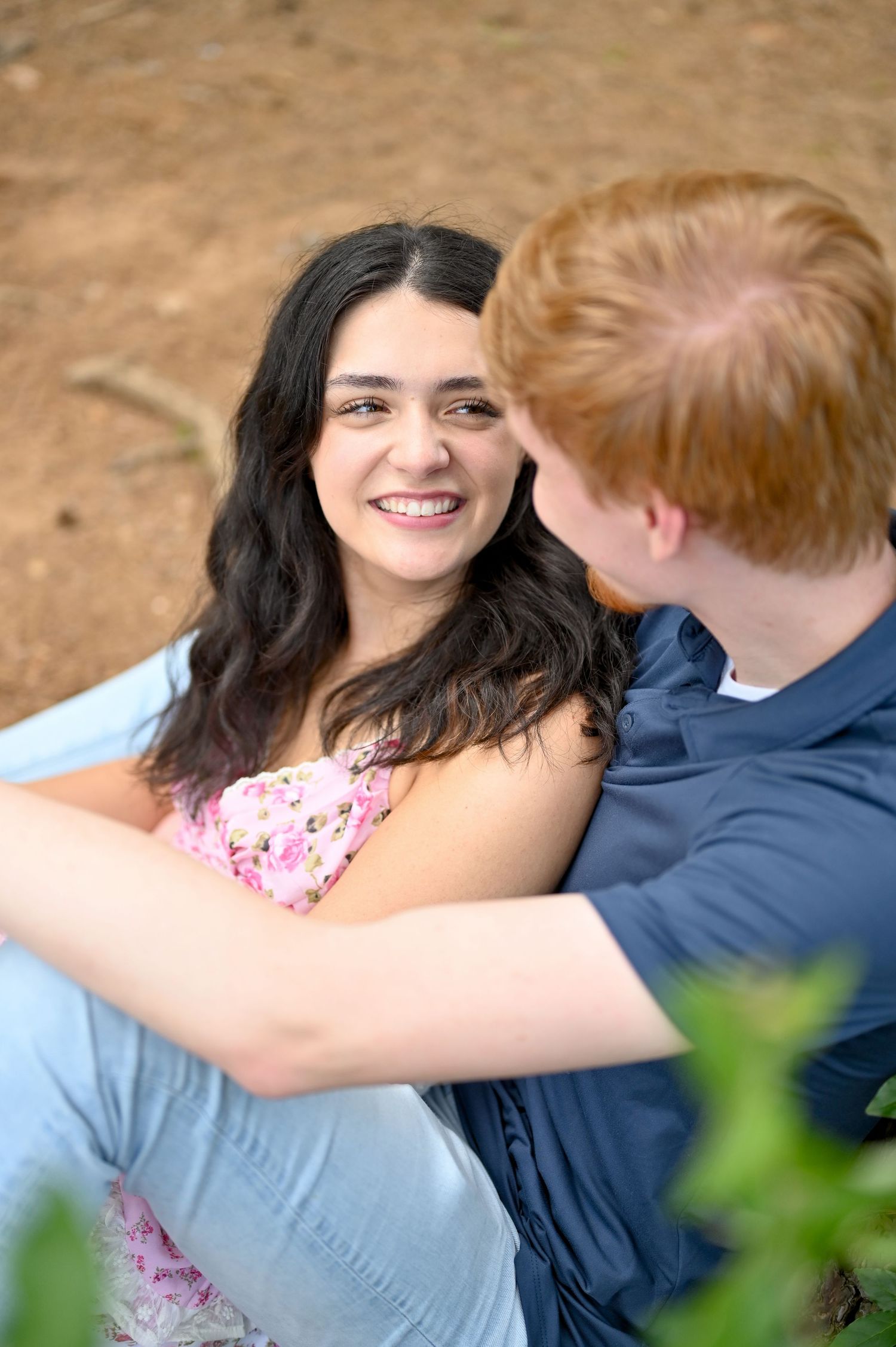 A smiling dark-haired woman sits outdoors with a red-haired man in a blue shirt during a couples photo session.