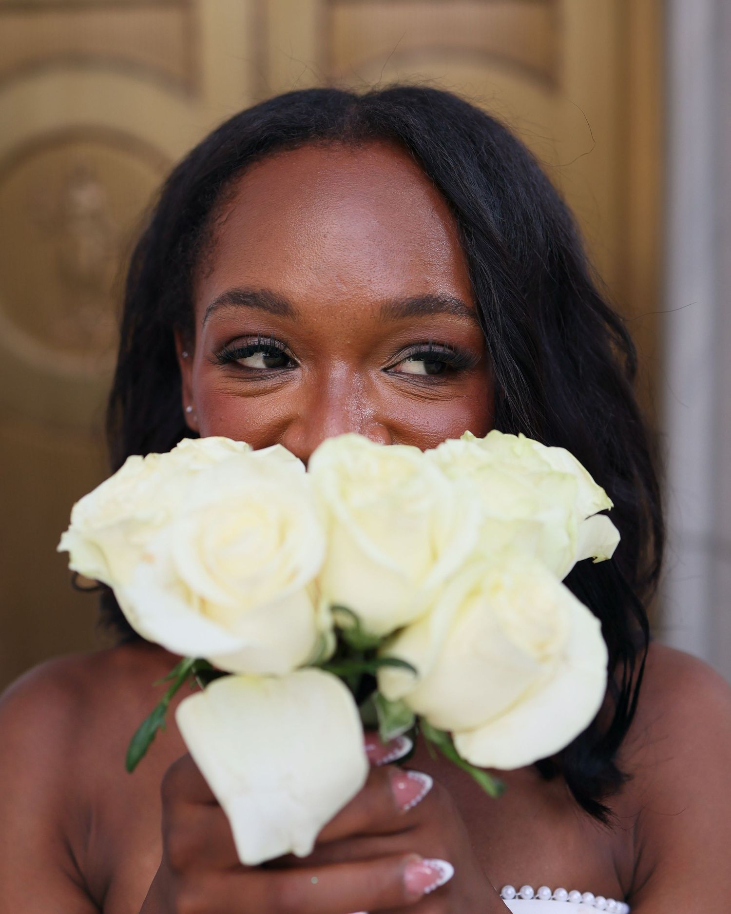 A Black woman holds white roses in front of her face, showcasing elegant makeup and pearl accessories.