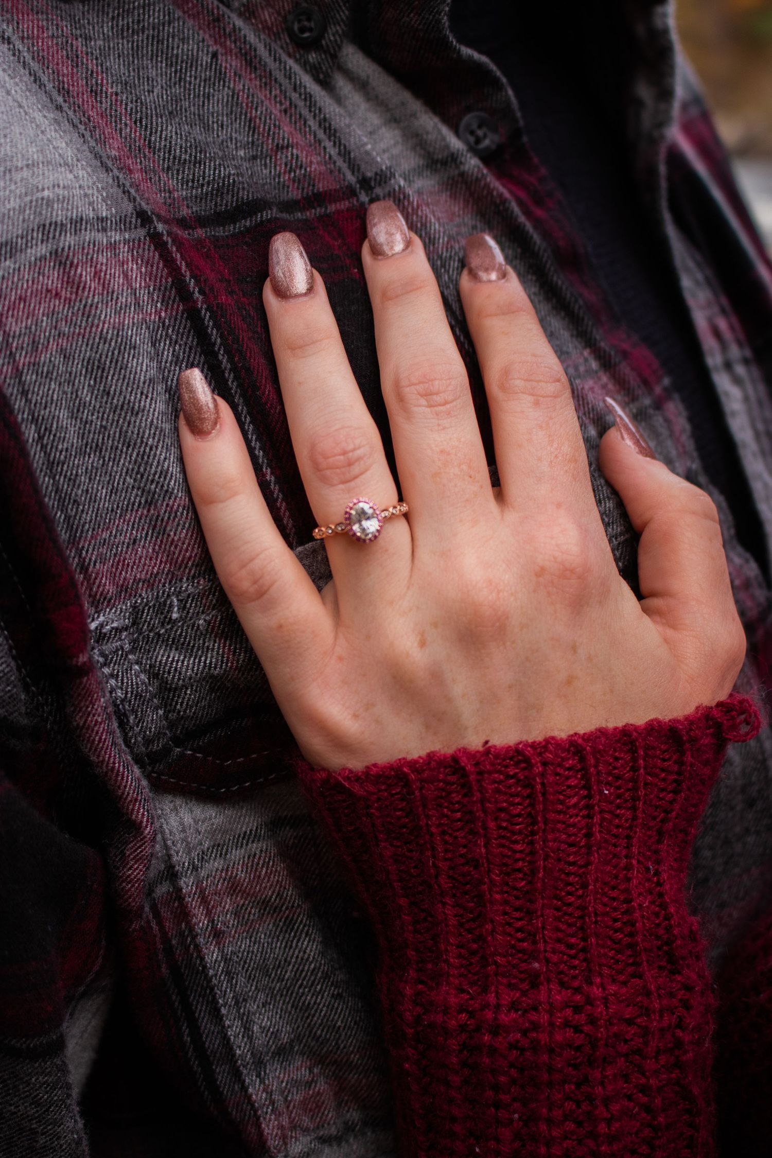 Close-up of a woman's hand wearing a rose gold diamond engagement ring, resting on a plaid flannel shirt.