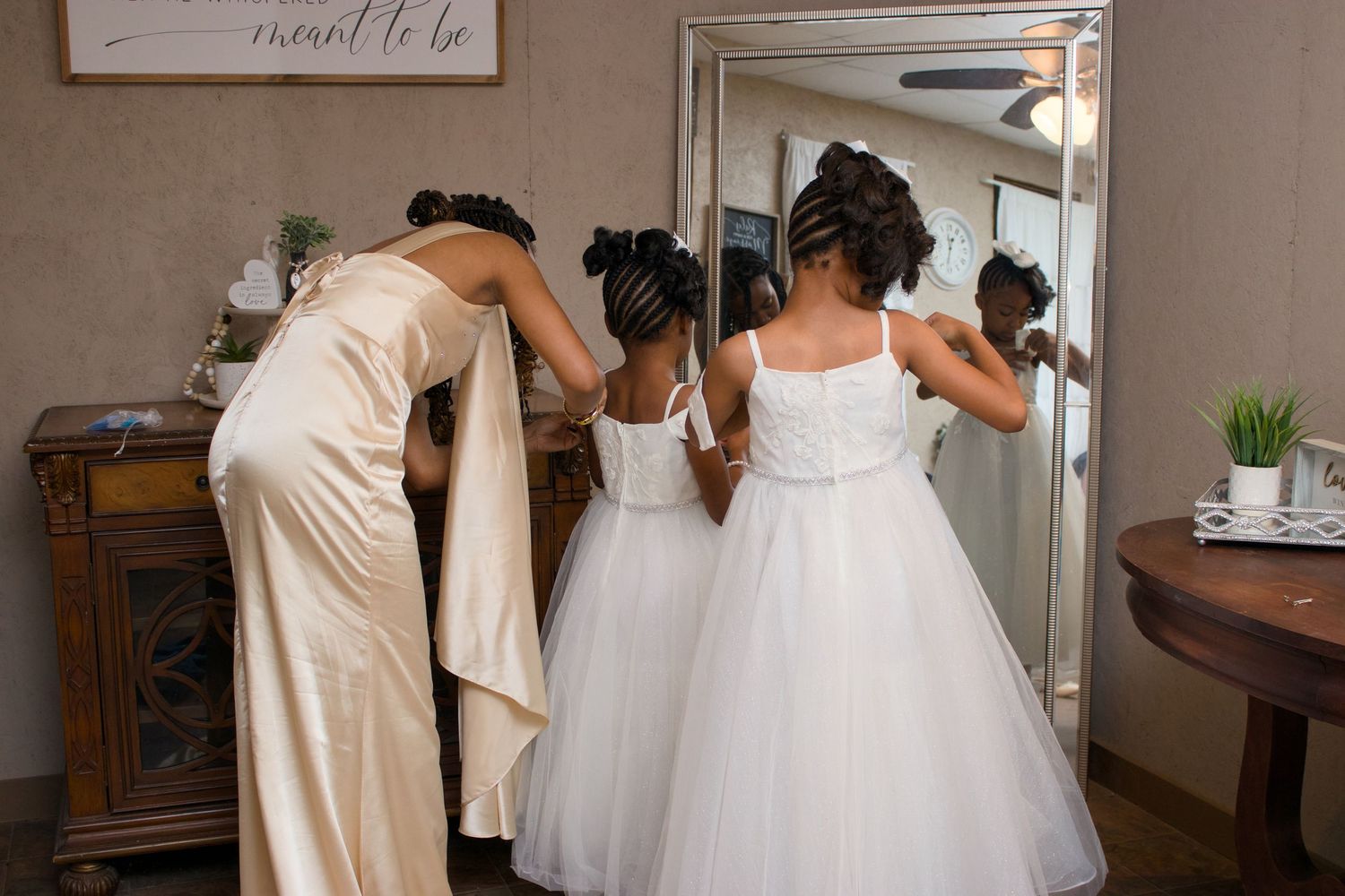 A woman in a champagne gown helps two young flower girls in white dresses prepare in front of a mirror.