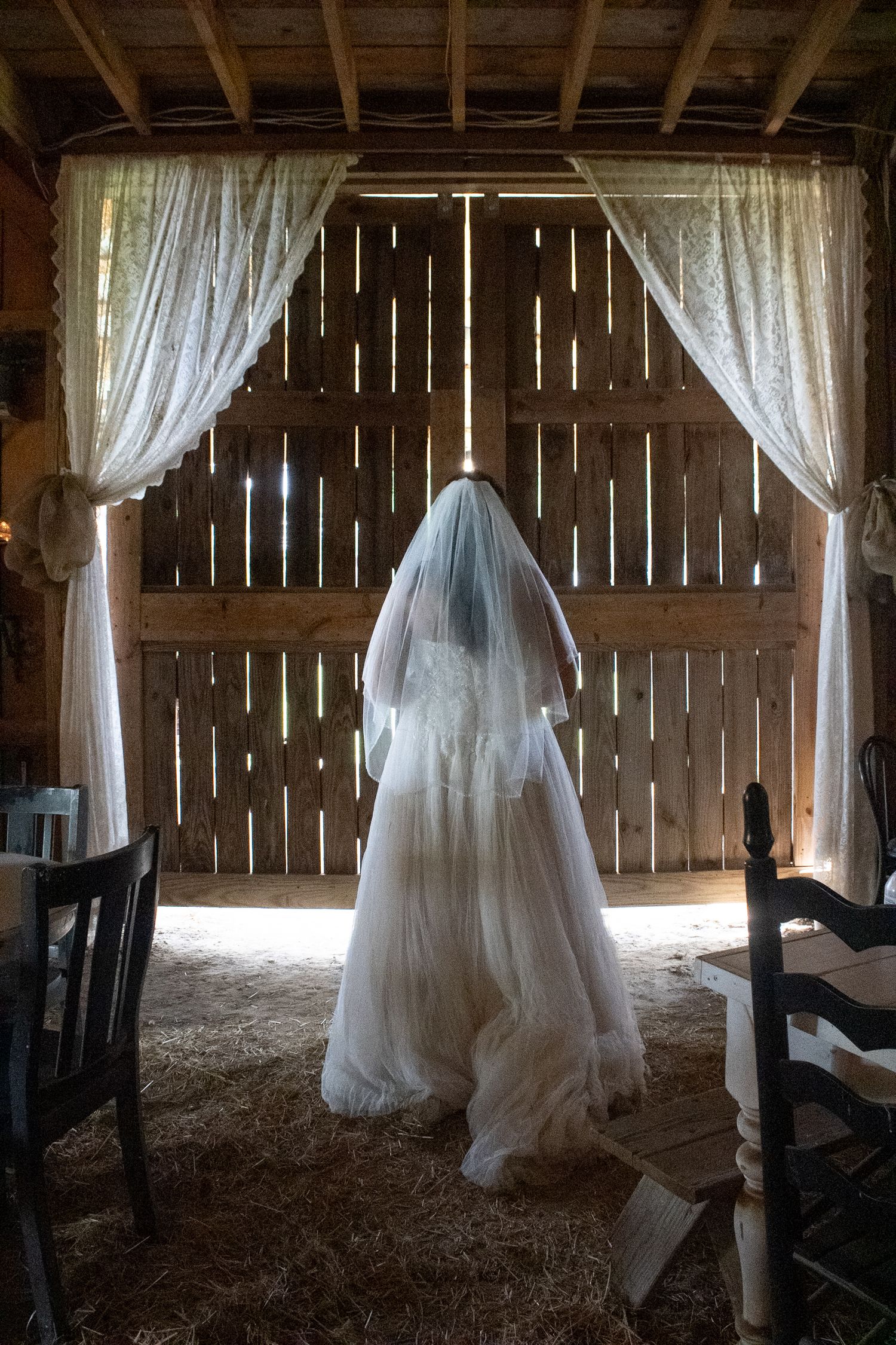 Bride in white wedding gown and veil standing in rustic barn doorway with sheer curtains and hay on floor.