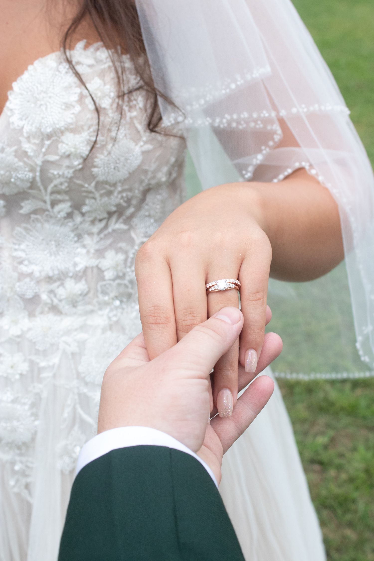 Groom in green suit holding bride's hand displaying diamond wedding ring on her finger during outdoor ceremony.