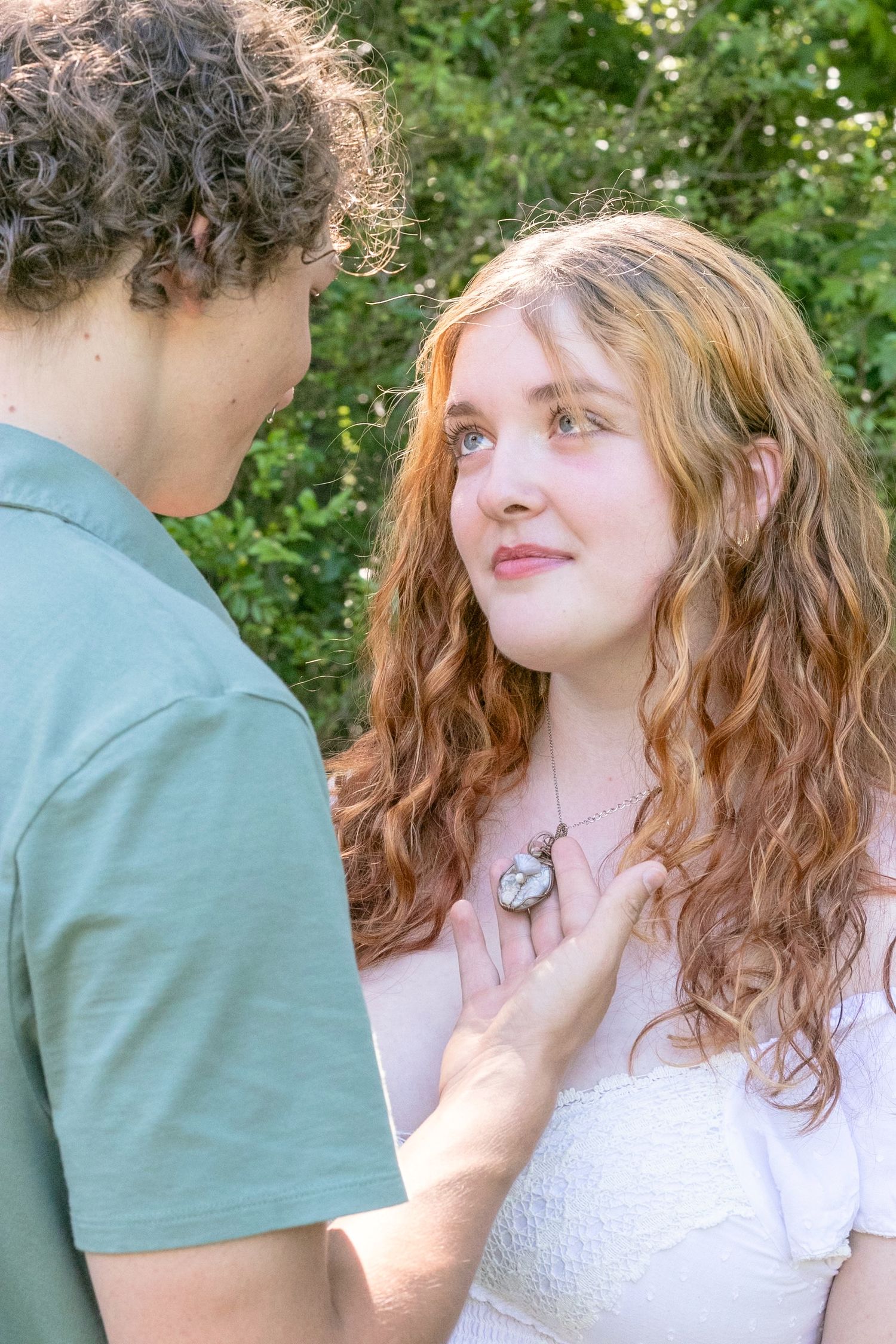 Young couple sharing a tender moment outdoors, redhead girl in white dress looking up at boy in green shirt.