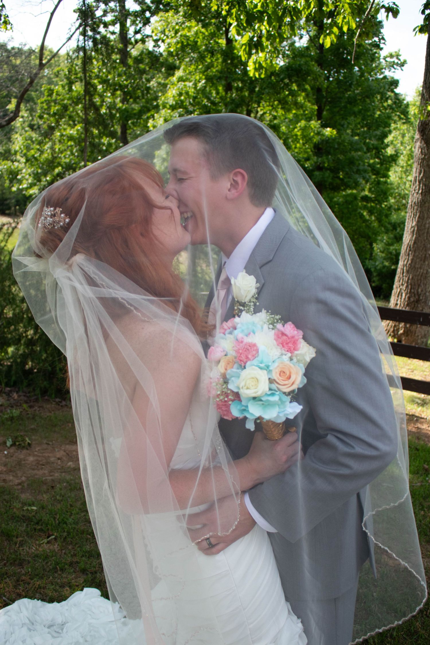 Bride and groom sharing romantic kiss under wedding veil outdoors with colorful bouquet.