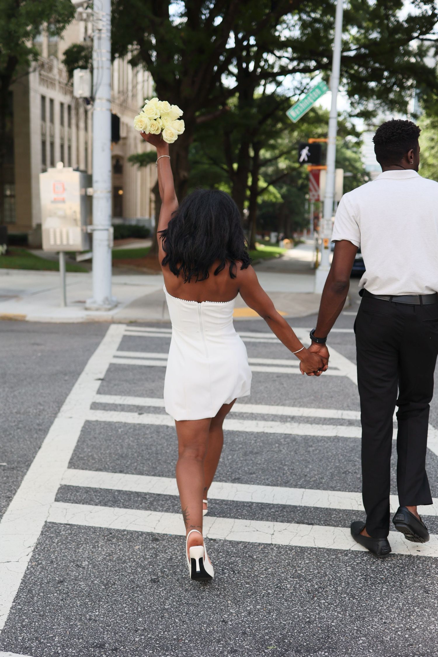 Newlywed Black couple holding hands crossing a street, bride in white mini dress raising bouquet.
