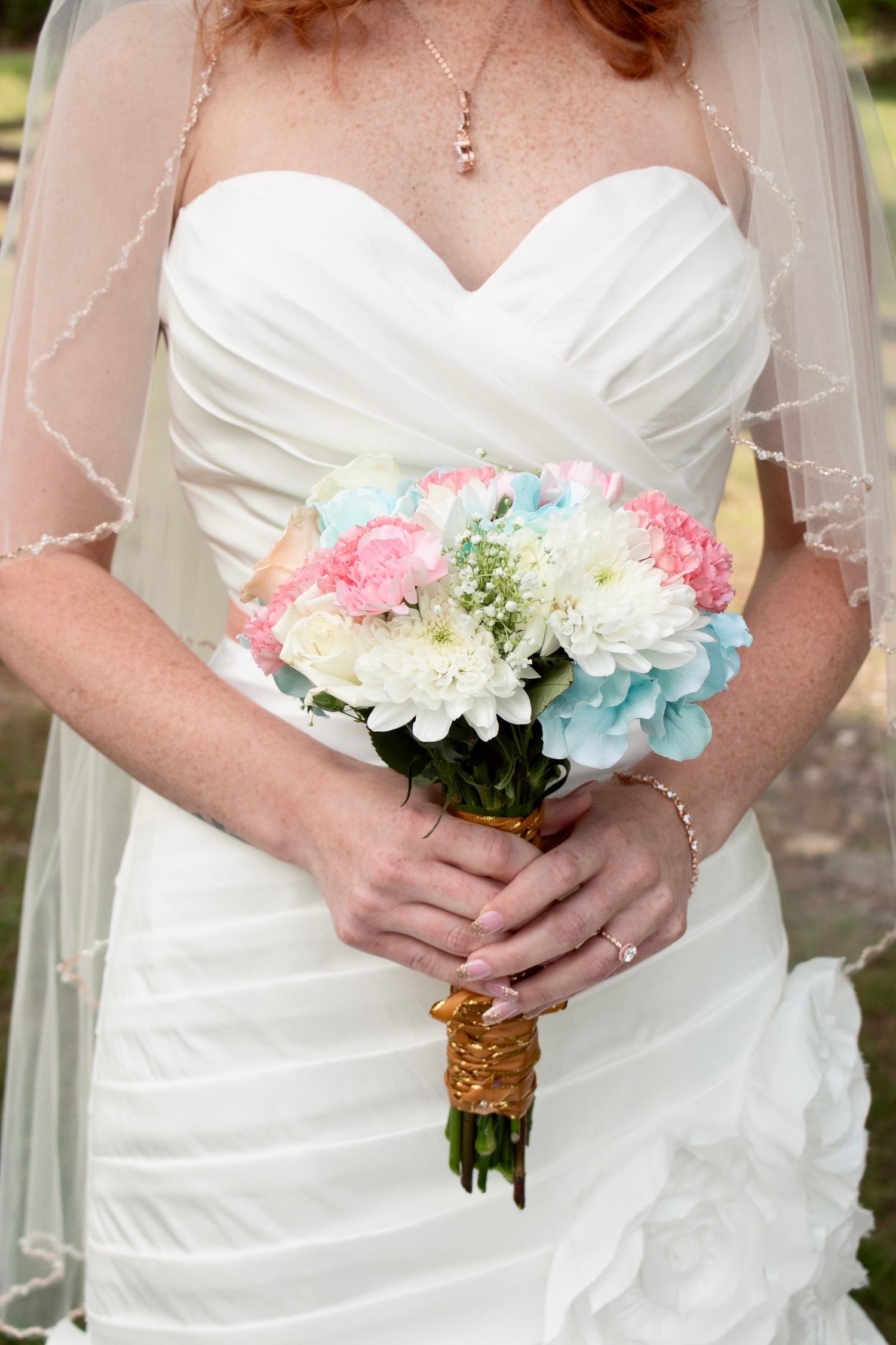 Bride in white strapless gown holding pastel bouquet of pink, blue, and white flowers with gold ribbon wrap.