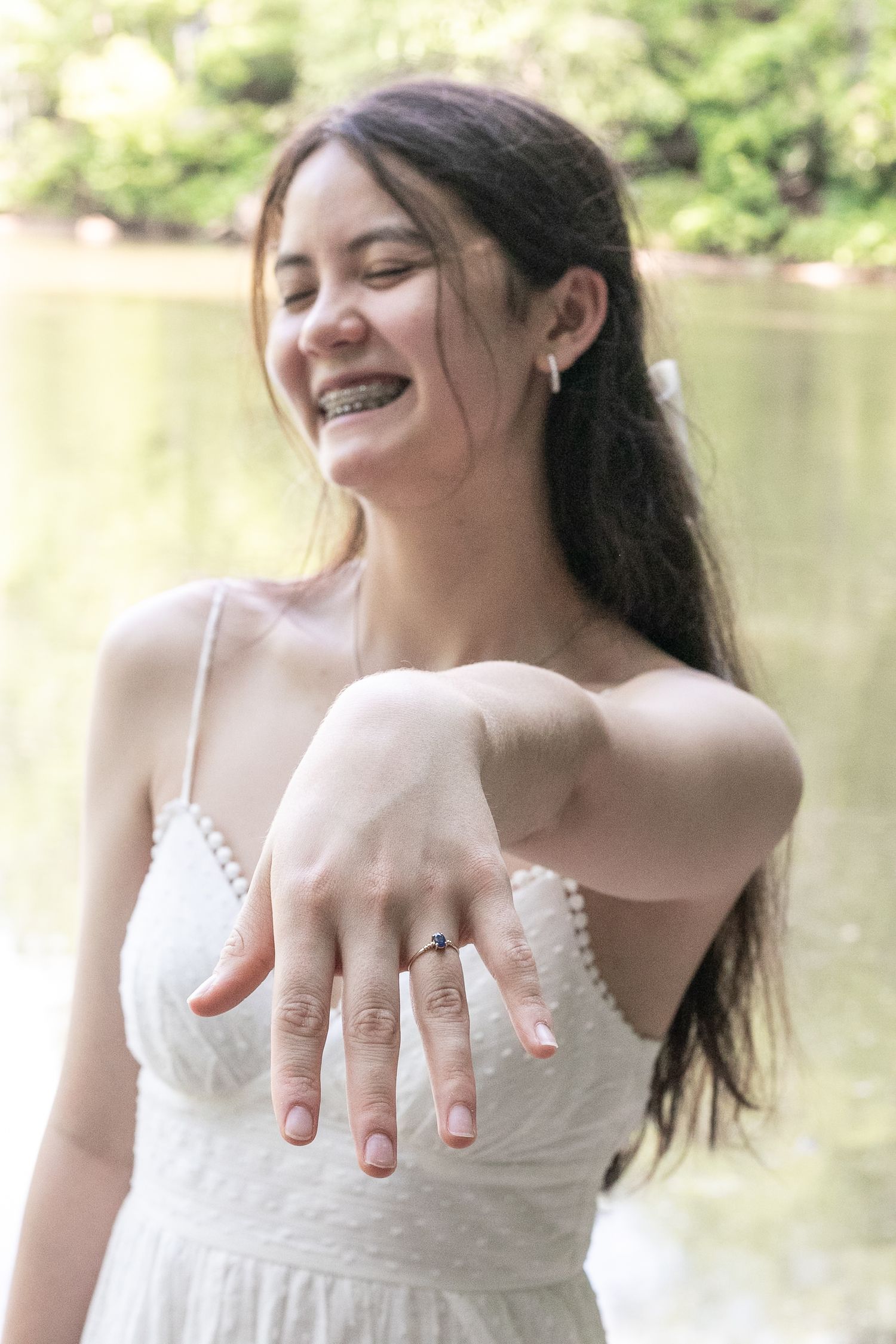 Smiling young woman in white dress showing off engagement ring outdoors near water.
