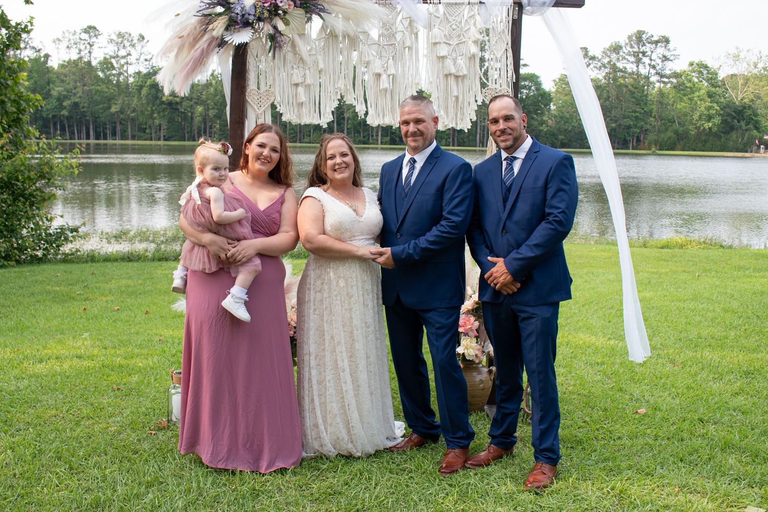 Wedding party poses outdoors near a lake with a boho arch, featuring a bride, groom, and guests in elegant attire.
