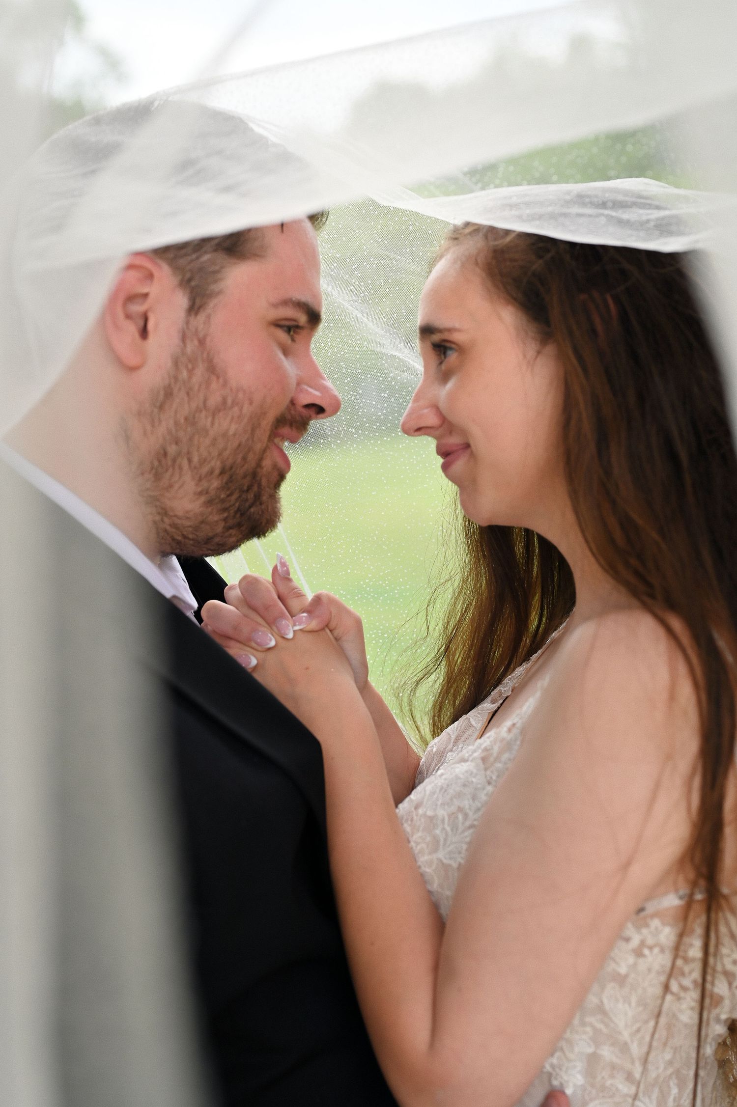 Bride and groom share an intimate moment under a wedding veil, holding hands and smiling at each other.