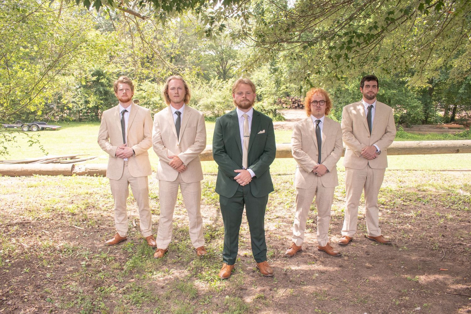 Groom in dark green suit stands with four groomsmen in tan suits outdoors under trees.