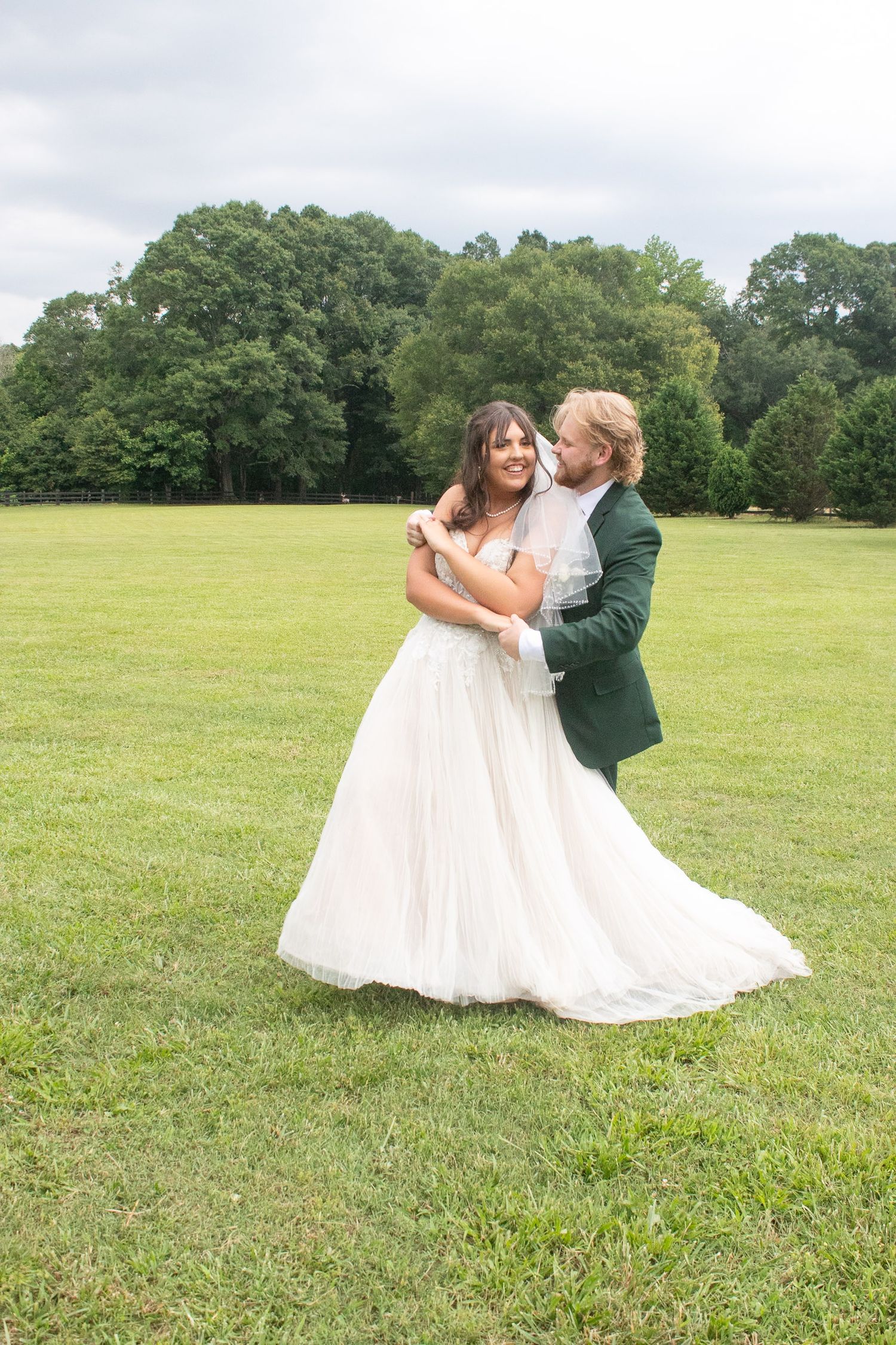 Bride and groom dancing joyfully on green lawn, groom in emerald suit kissing bride's cheek in white ballgown.