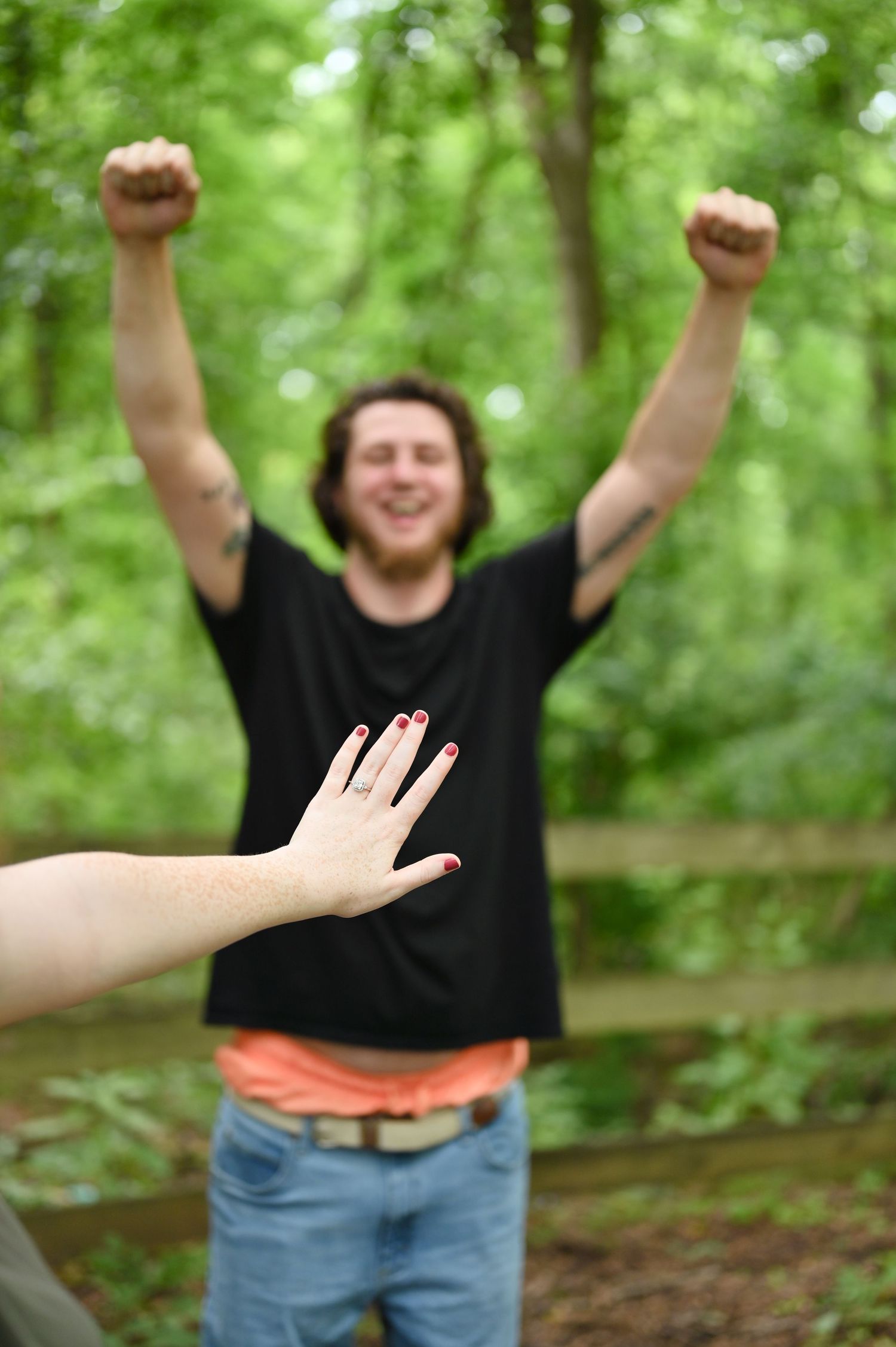 Man in black shirt cheering with raised fists outdoors as woman shows off engagement ring in foreground.