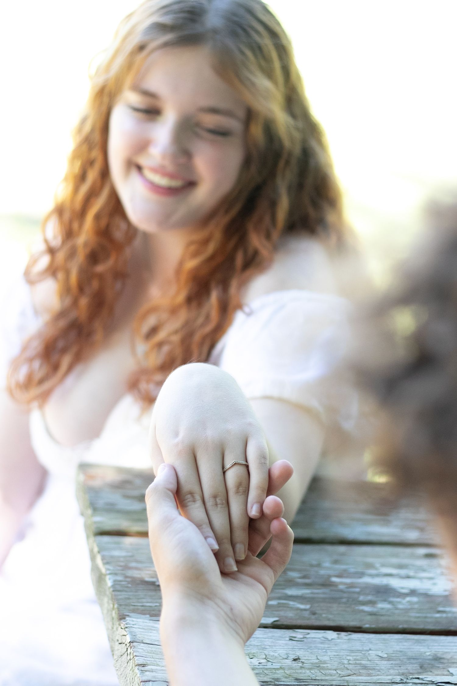 Smiling redhead bride in white dress showing off engagement ring while holding hands with partner on wooden dock.