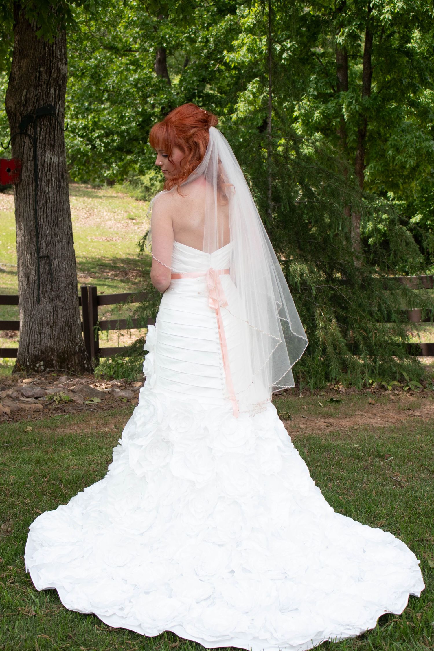 Bride in elegant white ruched wedding gown with veil and pink sash standing in garden.