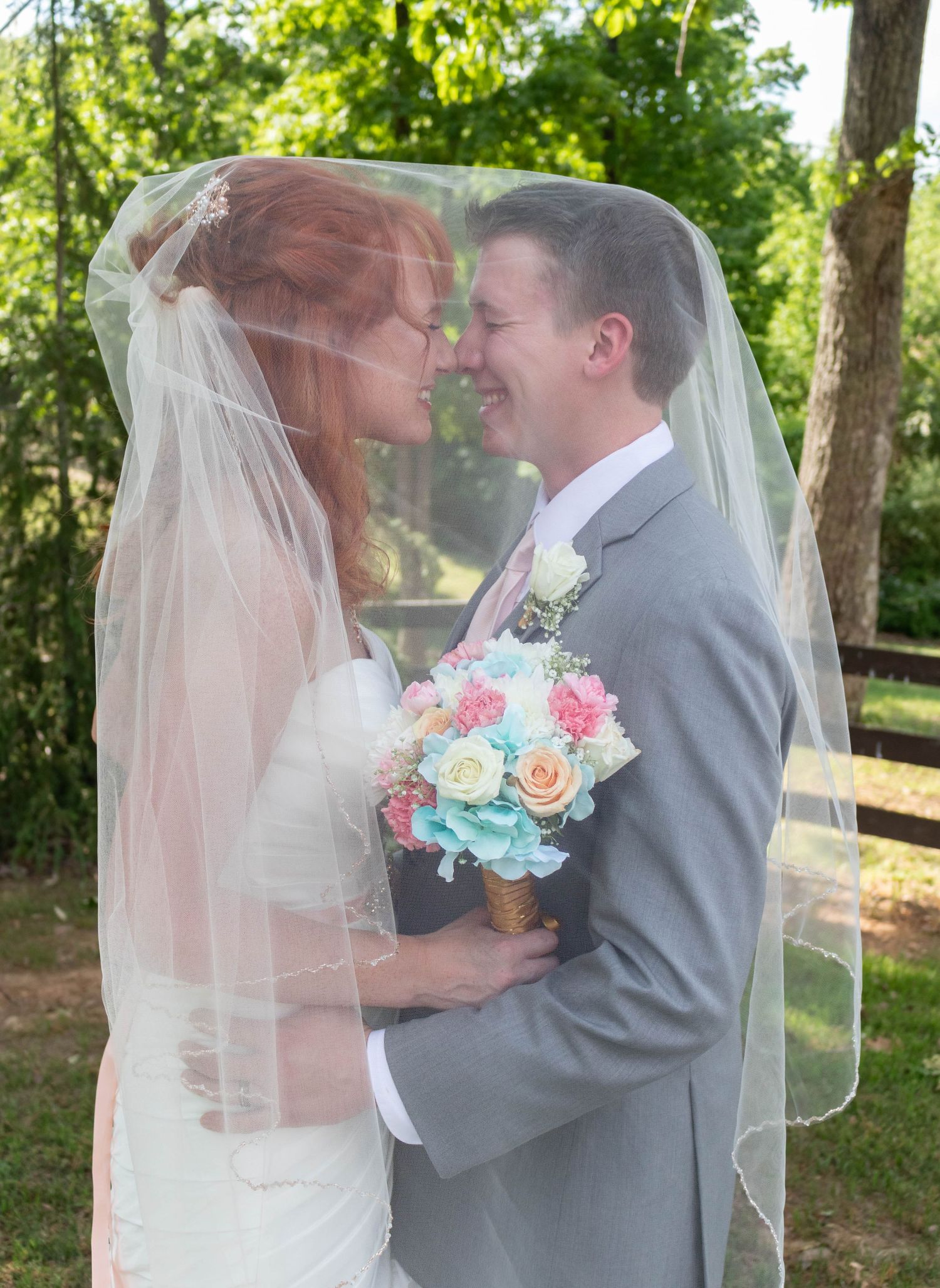 Bride and groom sharing intimate moment under wedding veil outdoors, bride holding colorful bouquet.